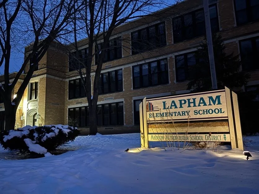 Snow-covered ground with a sign that reads "Lapham Elementary School" in front of a multi-story brick building at dusk.
