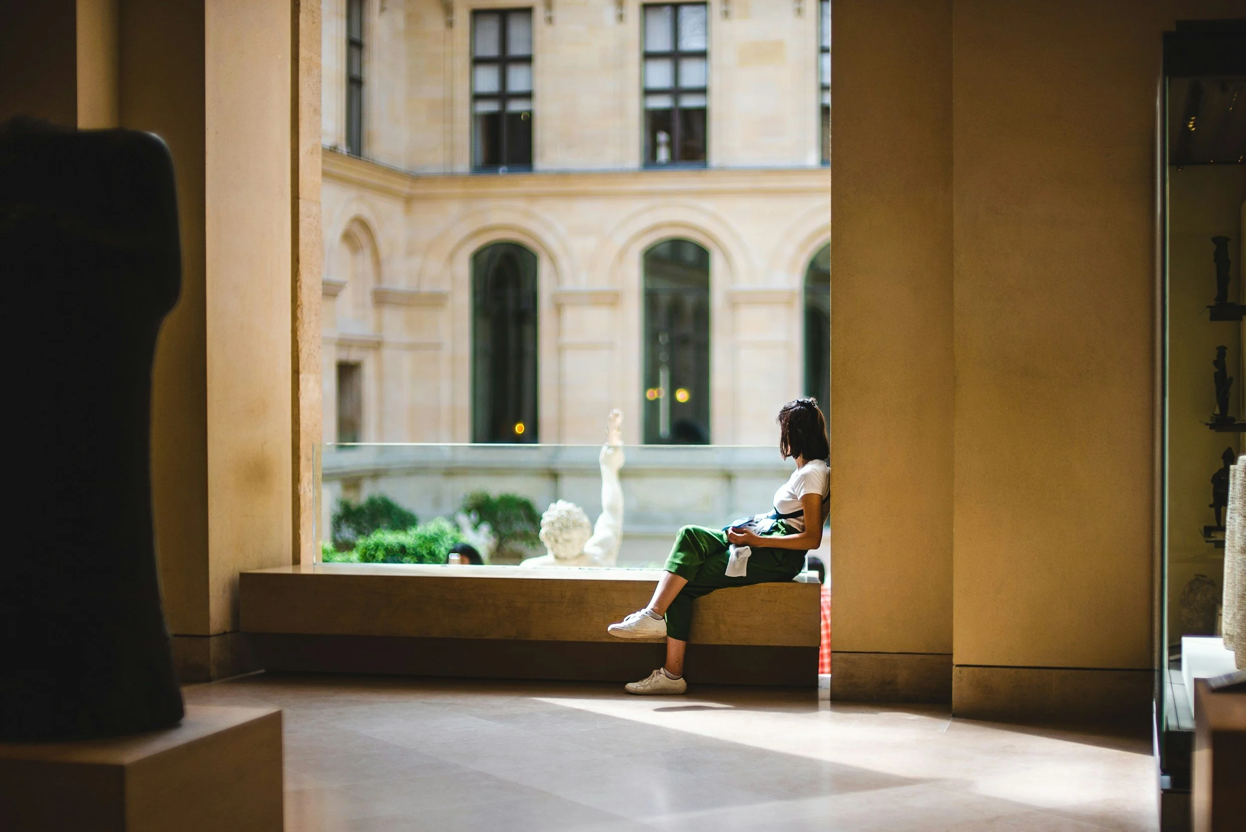 Woman sitting on a windowsill inside a building, looking outside at a courtyard with sculptures.