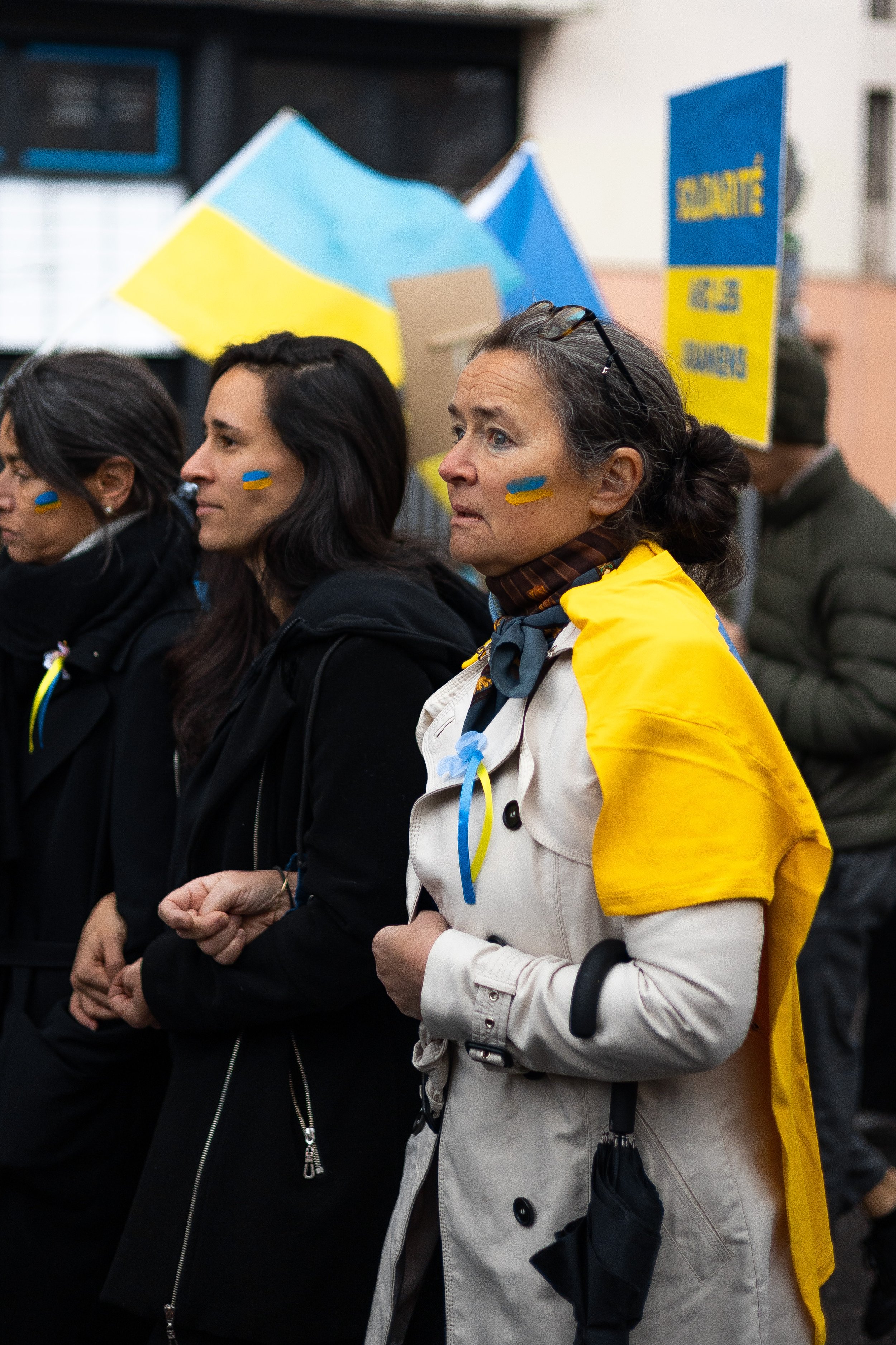 Des personnes participant à une manifestation avec des drapeaux ukrainiens. Elles portent des vêtements aux couleurs de l'Ukraine avec des accessoires bleus et jaunes. Une femme a un foulard jaune sur l'épaule et d'autres ont des autocollants de drap