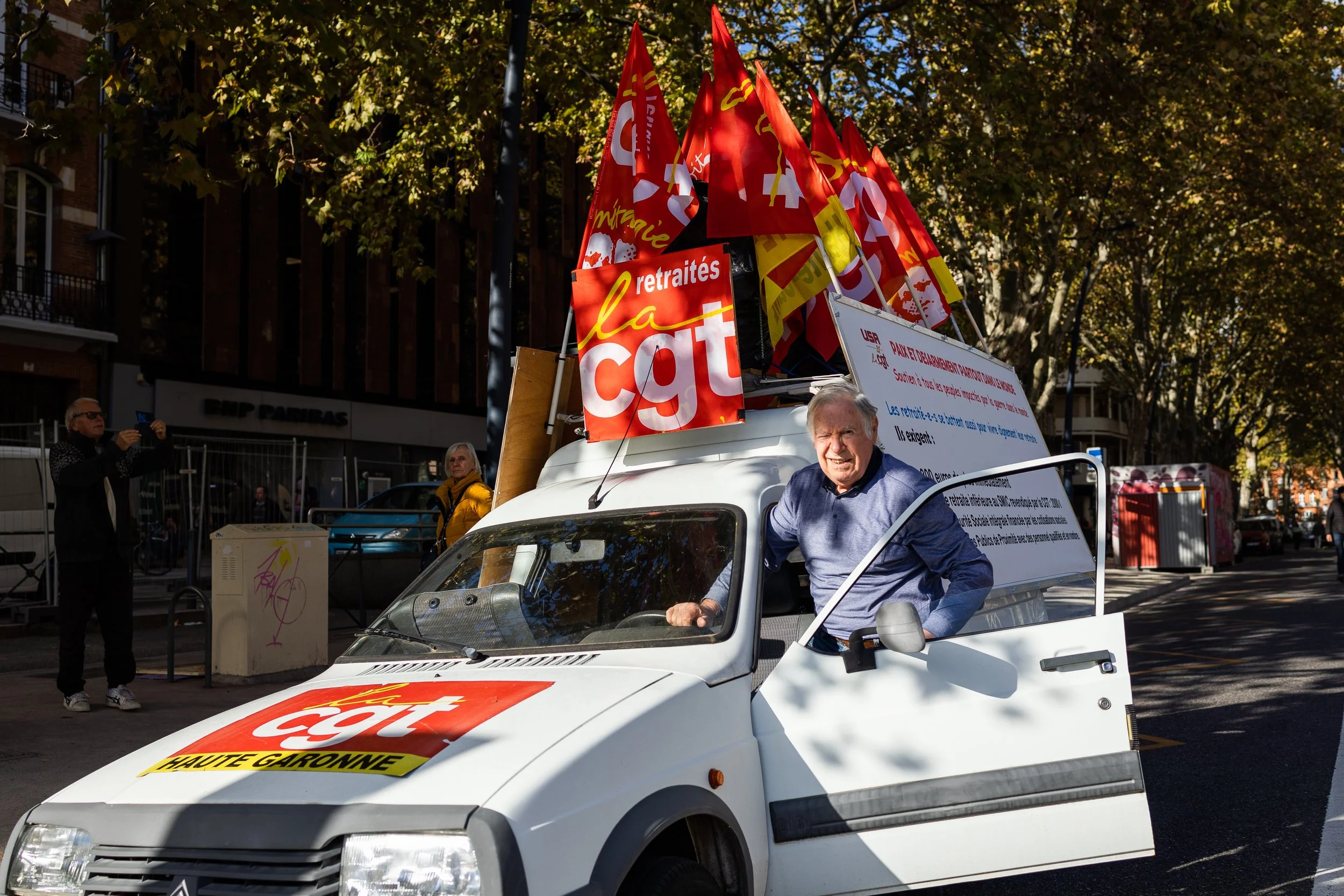 Voiture avec des drapeaux syndicaux CGT, manifestant assis, rue bordée d'arbres.