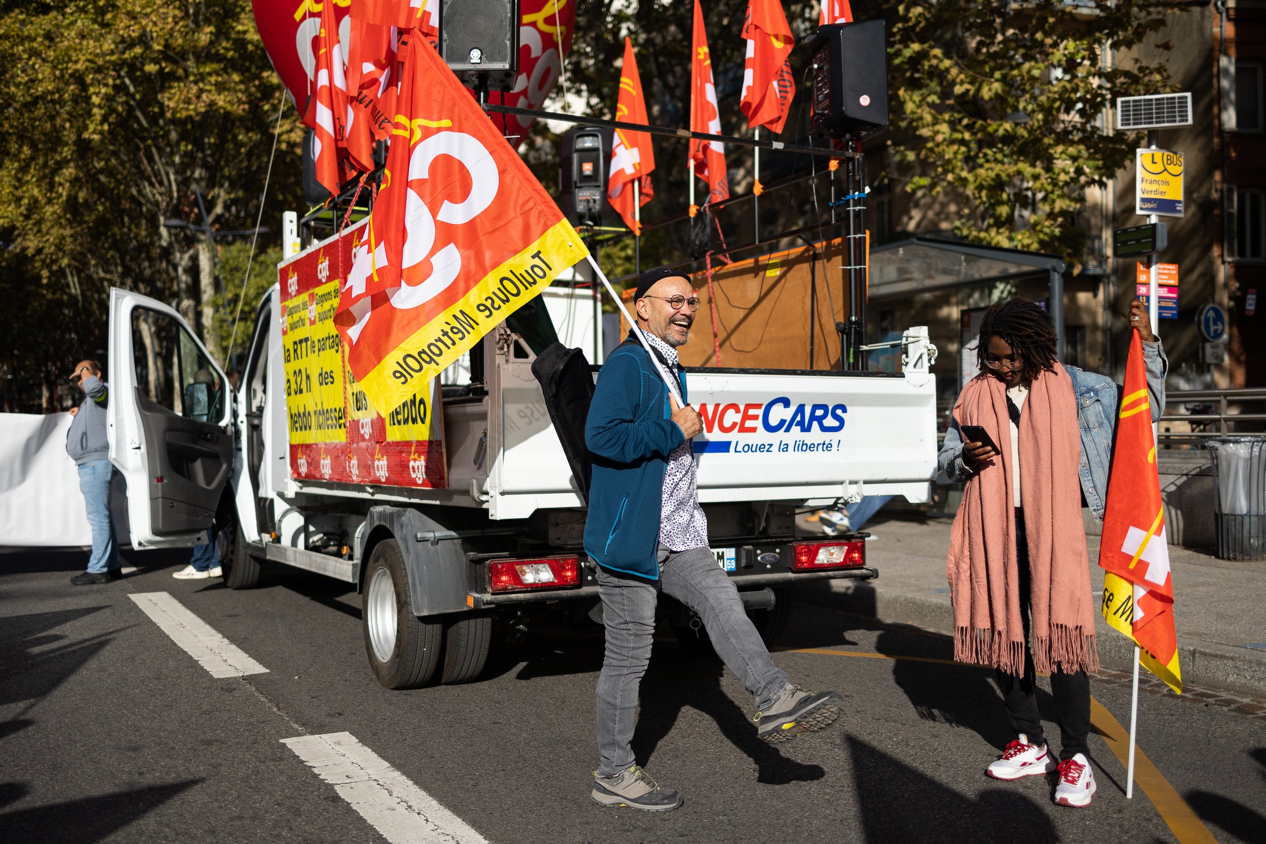 Manifestation avec camionnette de la CGT, drapeaux rouges, personnes souriantes tenant des drapeaux, rues de la ville en arrière-plan.