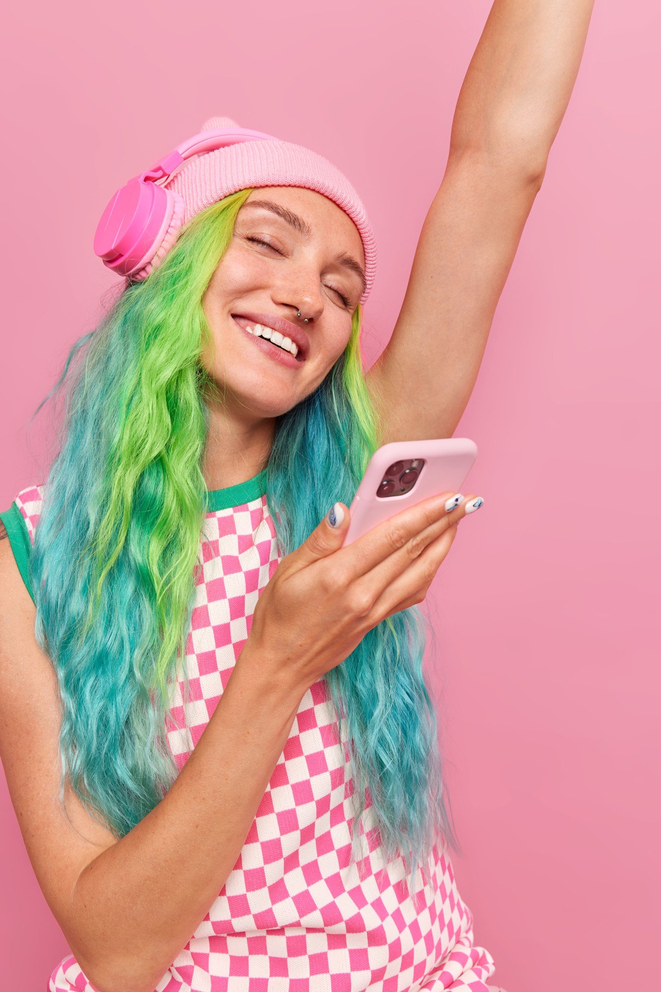 Young woman with rainbow-colored hair, pink beanie, and pink headphones smiling while looking at her smartphone against a pink background.