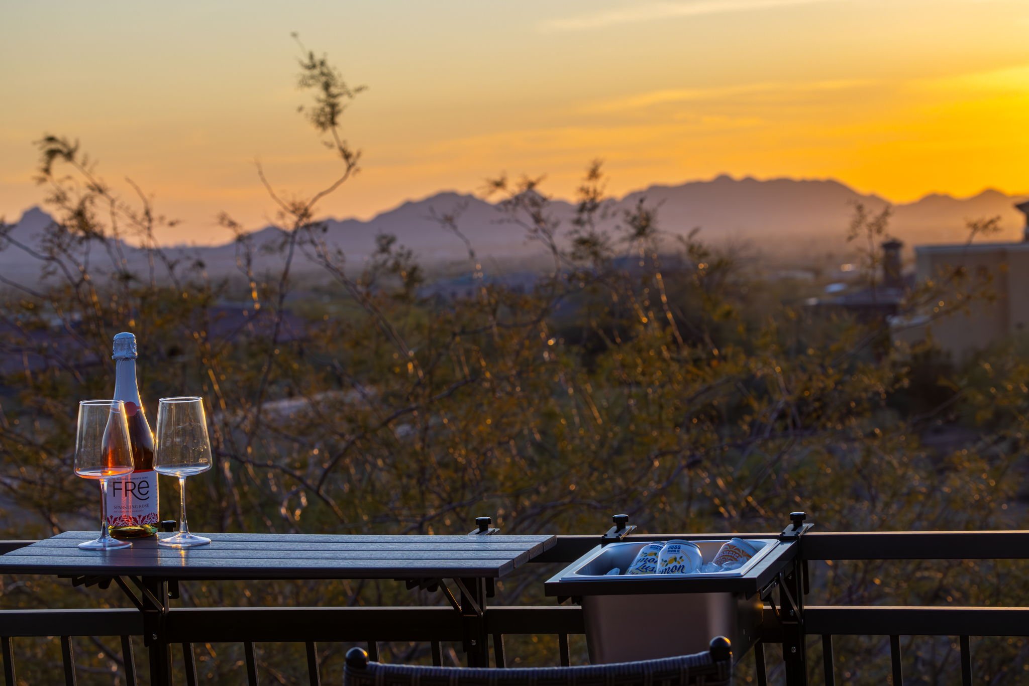 A balcony at sunset with a bottle of rosé wine and two glasses, and a cooler with coated cans, against a background of mountains and trees.