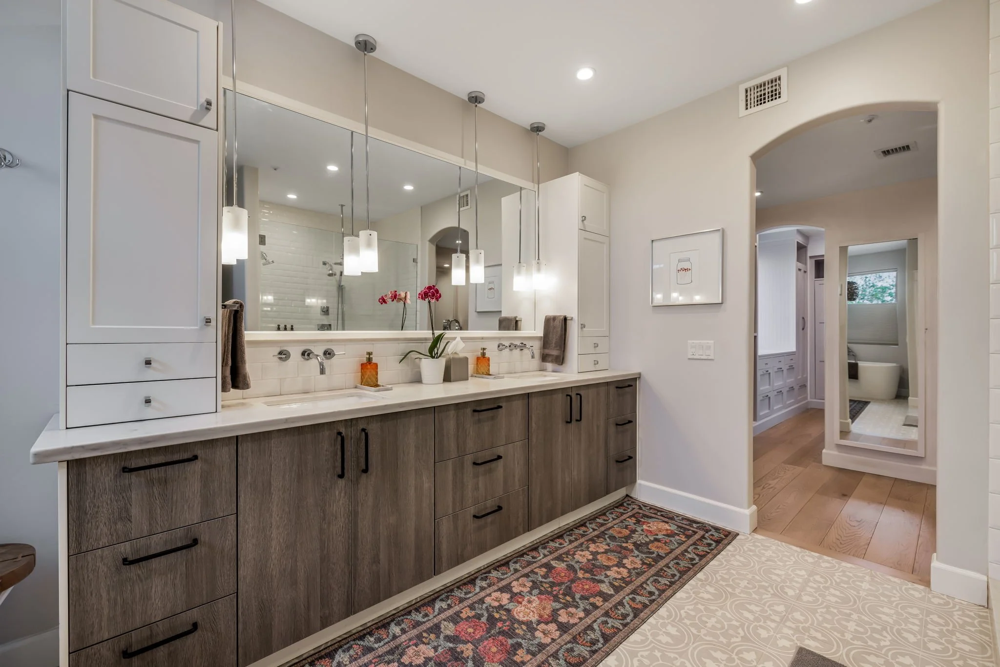 Modern bathroom with a large mirror, pendant lights, a double sink vanity with wooden cabinets, a decorative orchid, and a view into the shower area, with a separate toilet room visible through an open doorway.