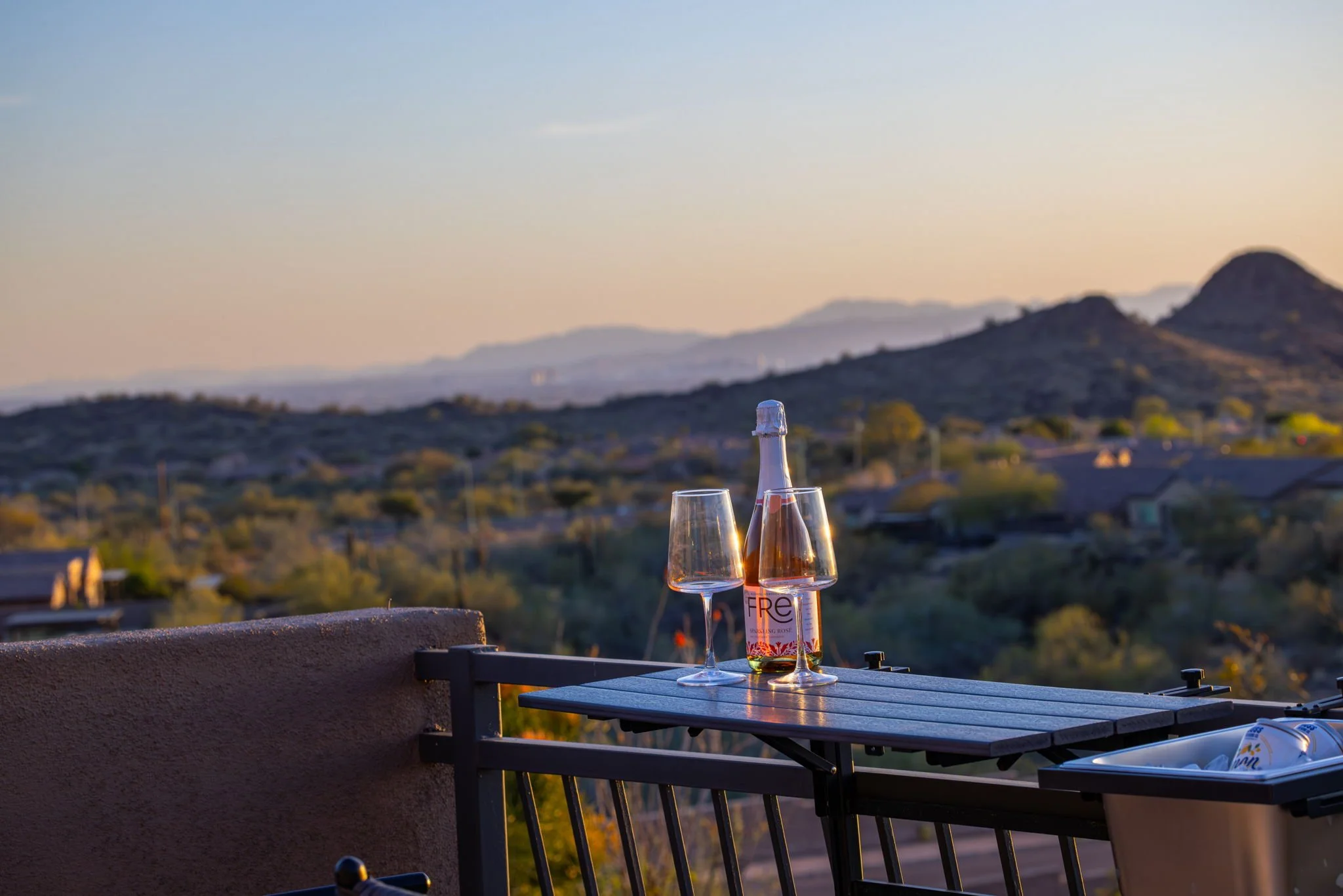 A table on a balcony holds a bottle of rosé wine and two empty wine glasses, with a scenic view of rolling hills and houses in the background at sunset.