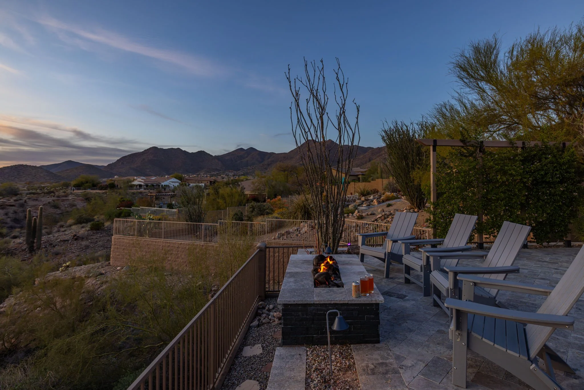 Outdoor patio with four Adirondack chairs facing a fire pit, desert landscape, mountains in the background, trees, and a sunset sky.