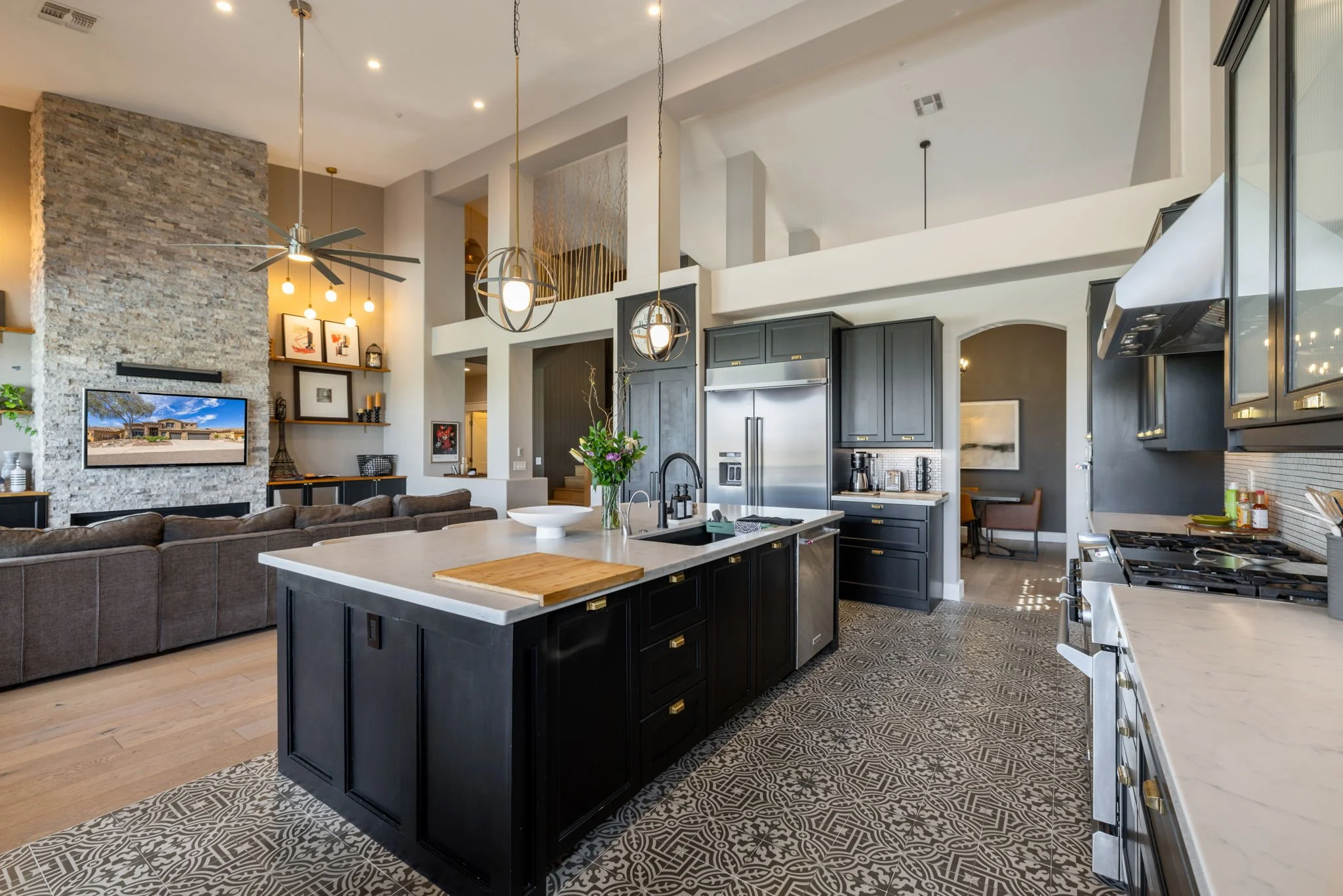 Open-concept kitchen and living room with black kitchen cabinets, a kitchen island with a white countertop, stainless steel refrigerator, and decorative hanging lights, living room features a stone fireplace, a large couch, and a wall-mounted TV.