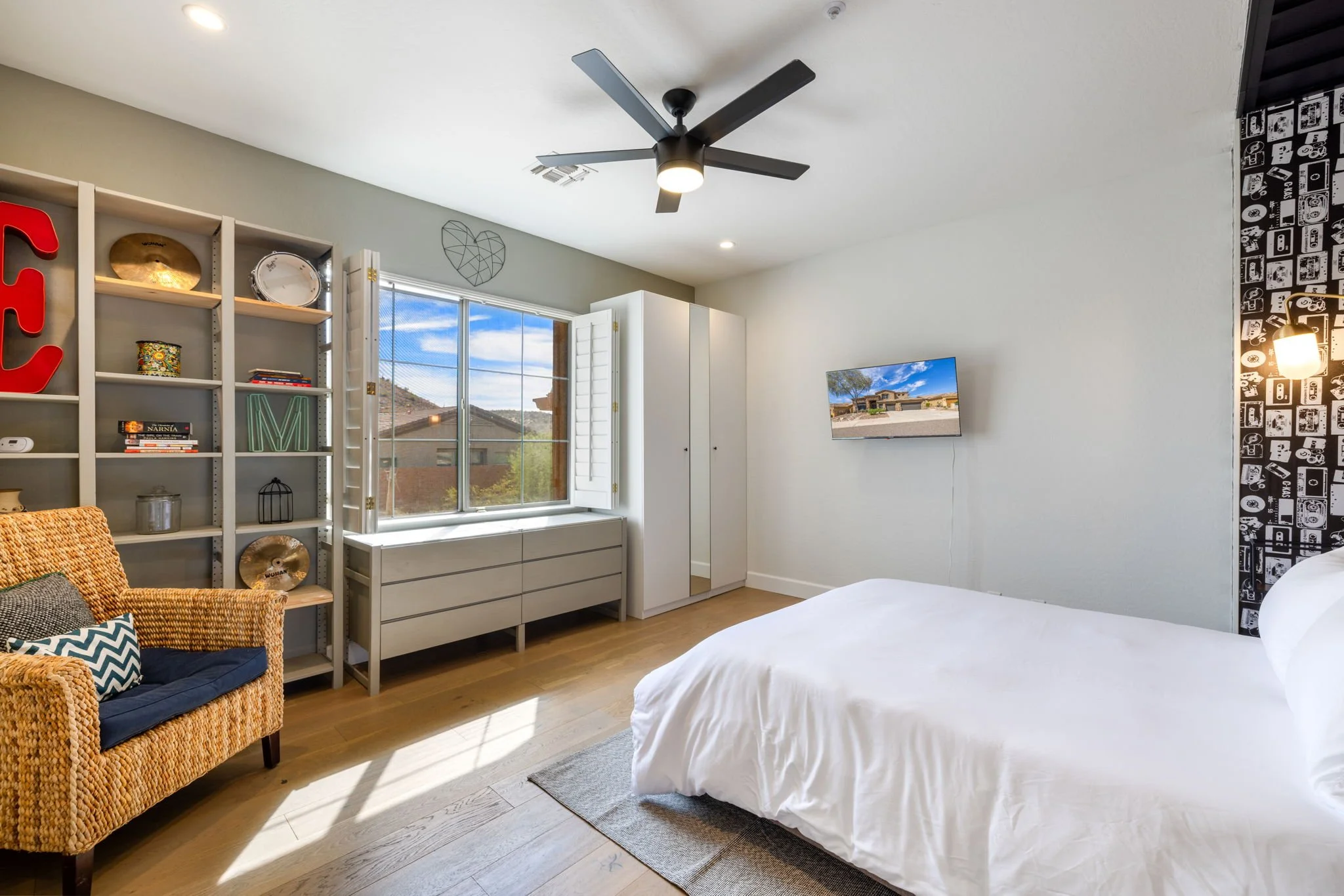 Modern bedroom with white walls, a bed with white bedding, a window with mountain views, a gray shelving unit with decor, a wicker chair with cushions, a wall-mounted TV, a ceiling fan, and patterned wallpaper on one wall.