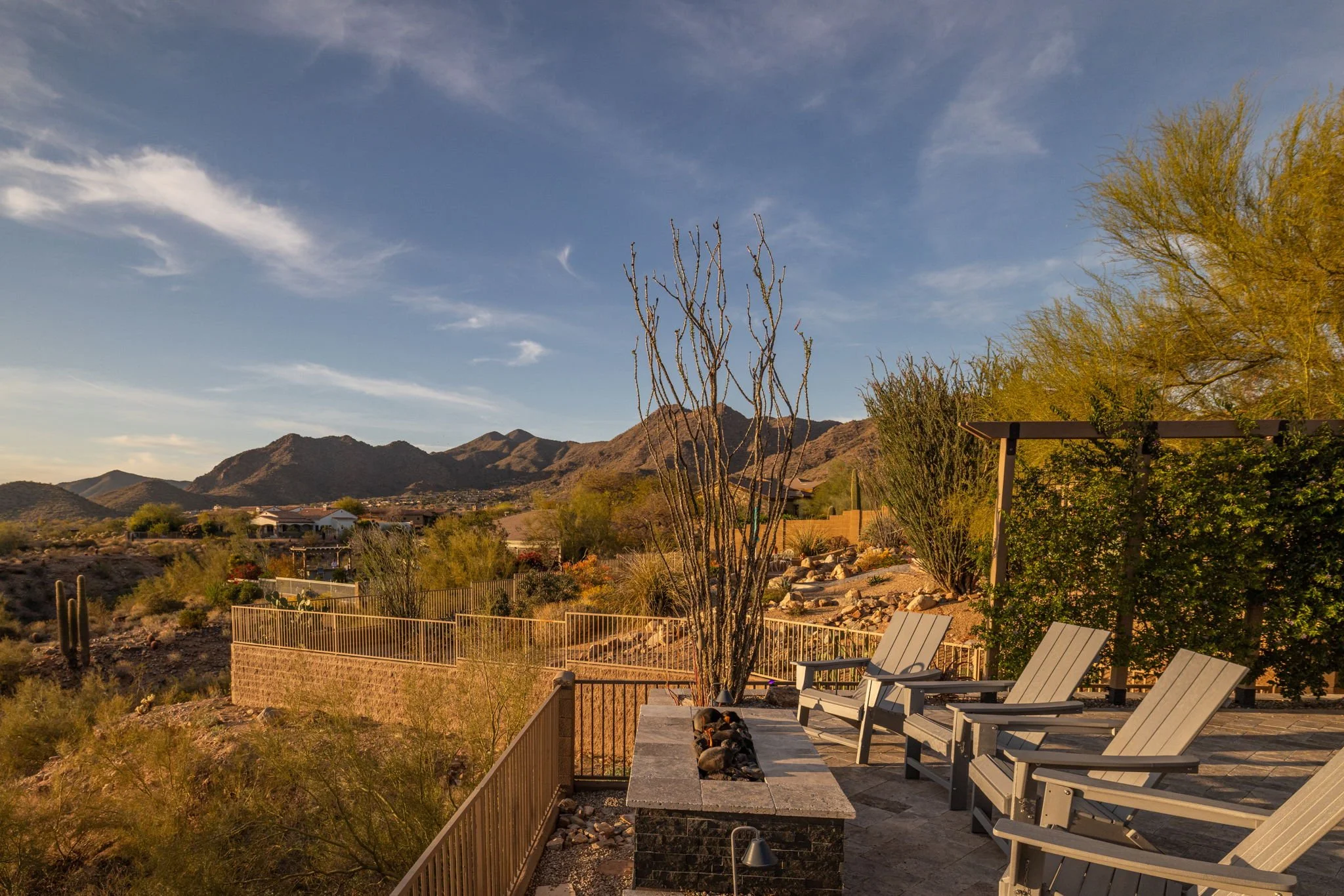 A desert landscape with mountains in the background, featuring a patio with three Adirondack chairs, a tall leafless cactus, and various desert plants, under a blue sky with some clouds.