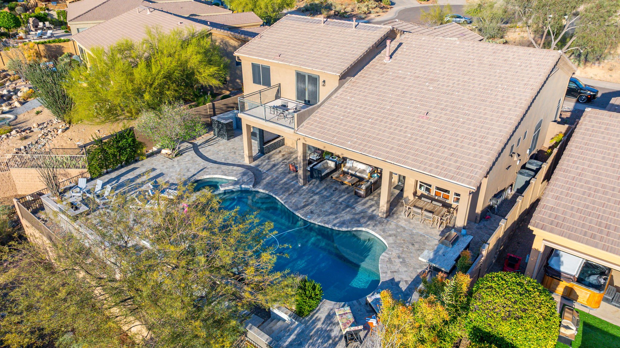Aerial view of a backyard with a kidney-shaped swimming pool, patio furniture, and a small hot tub, attached to a two-story house with beige exterior walls and a tiled roof, surrounded by trees and neighboring houses.