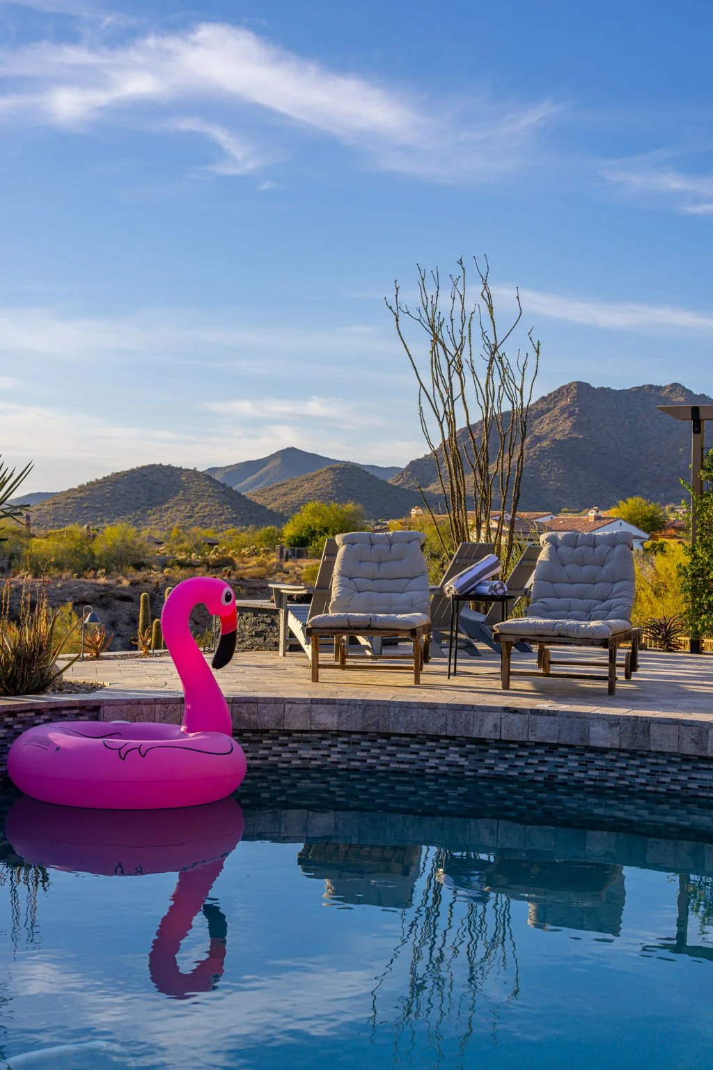 A backyard pool with a pink flamingo inflatable, two lounge chairs with cushions, and scenic mountains in the background under a blue sky with wispy clouds.