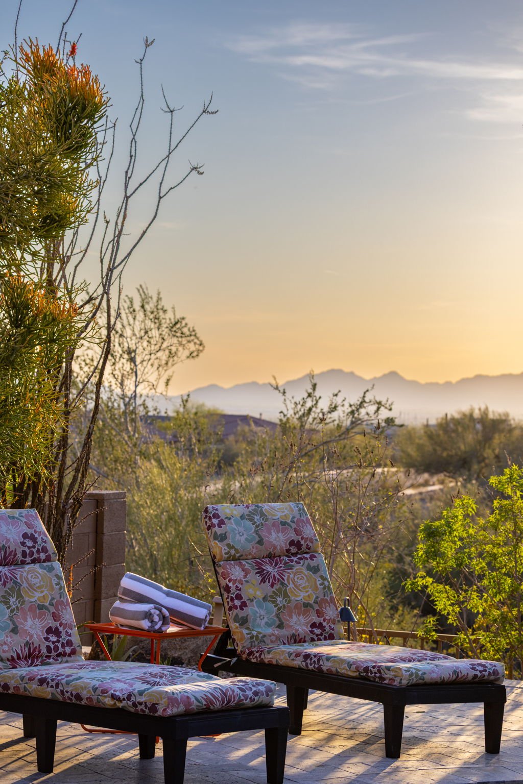 Two lounge chairs with floral cushions on a patio, with folded towels on a small orange table, overlooking a desert landscape with mountains at sunset.