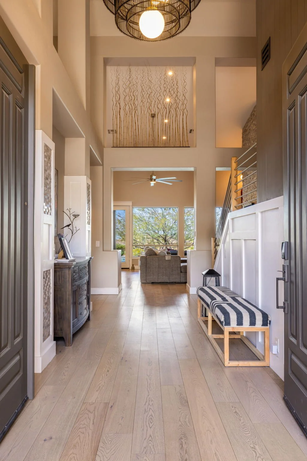 Interior view of a modern home entryway with light wood flooring, a black and white striped bench, and decorative lighting fixtures, leading to a living room with large windows and a view of trees outside.