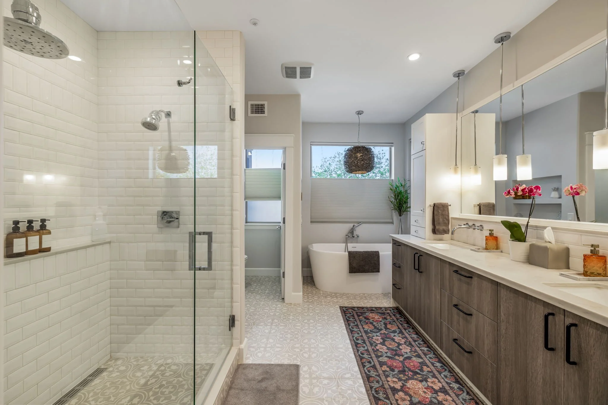 A modern bathroom with a glass-enclosed shower, a bathtub near large window, a long vanity with a large mirror, decorative lighting, and various bathroom accessories.