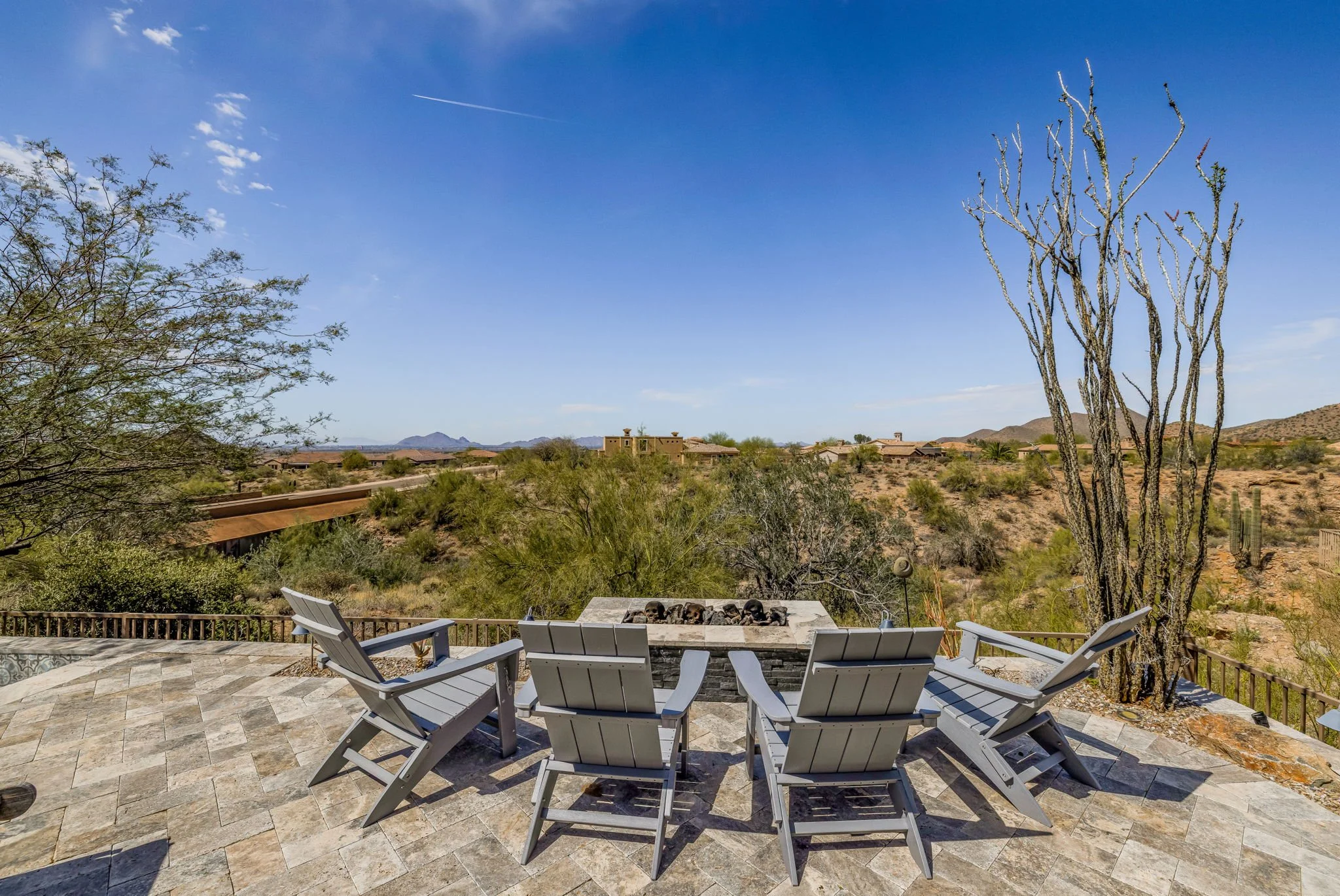 Scenic desert patio with six gray Adirondack chairs arranged around a fire pit, overlooking a desert landscape with cacti, bushes, and mountains under a blue sky.
