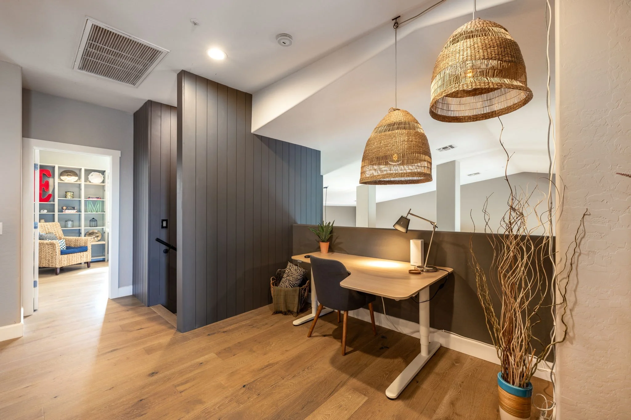 Home office corner with a wooden desk, black ergonomic chair, desk lamp, potted plant, decorative dried branches in a blue and tan vase, hanging woven pendant lights, dark wood slatted wall, and a partition wall.