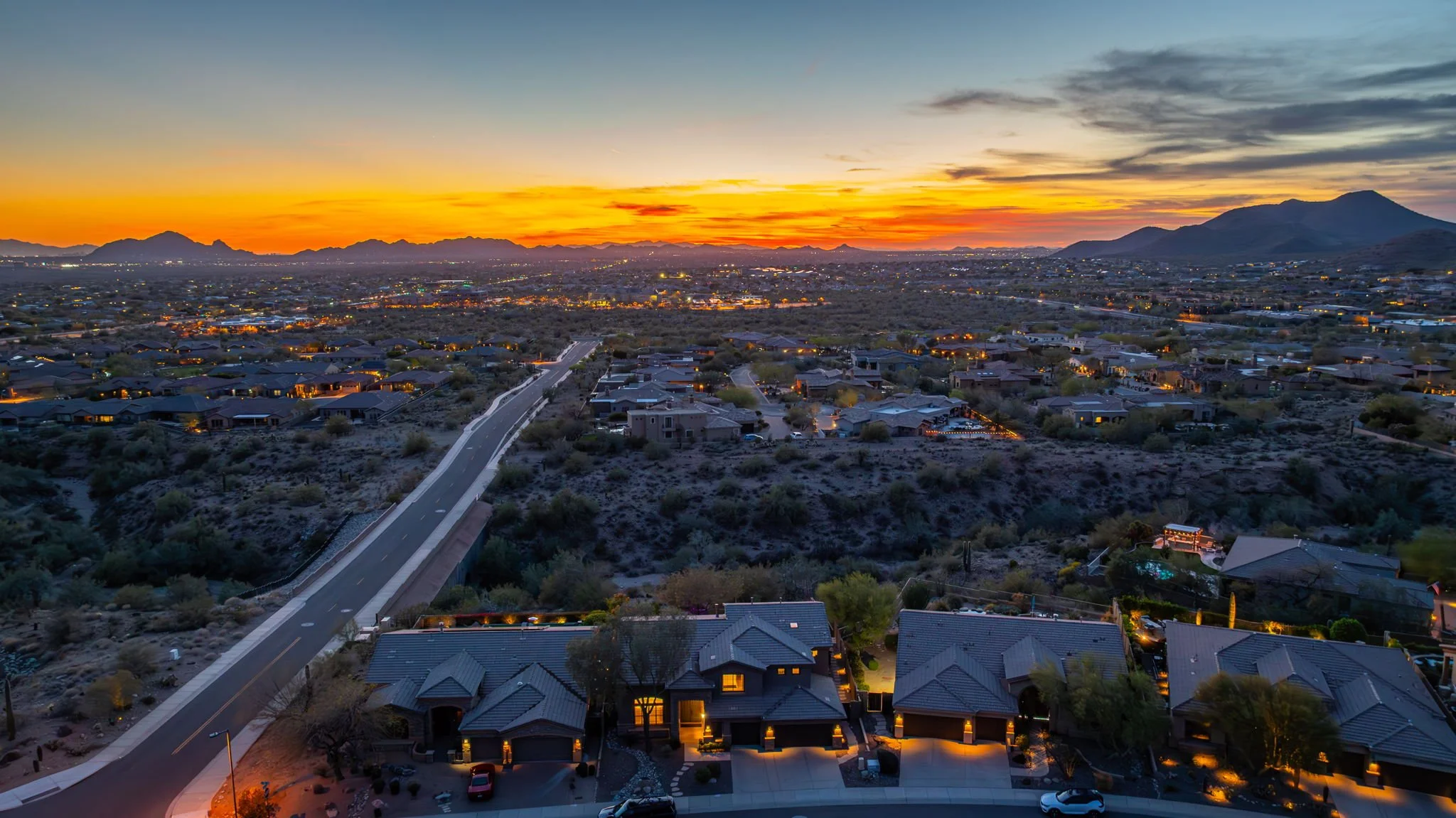 Aerial view of a suburban neighborhood at sunset, with houses, a winding road, and mountains in the distance.