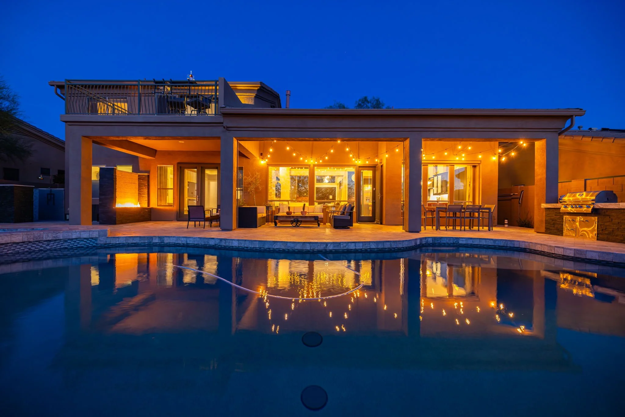 Backyard with a swimming pool, illuminated by warm string lights, showing a house with an outdoor patio and furniture at dusk.