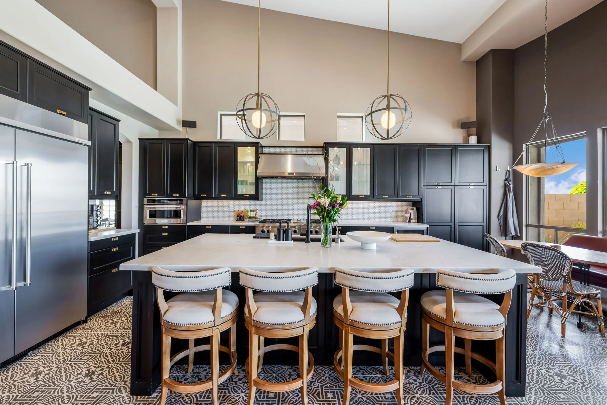 Modern kitchen with black cabinetry, large white island, stainless steel appliances, pendant lights, patterned tile floor, and windows with a view of a brick wall and trees.