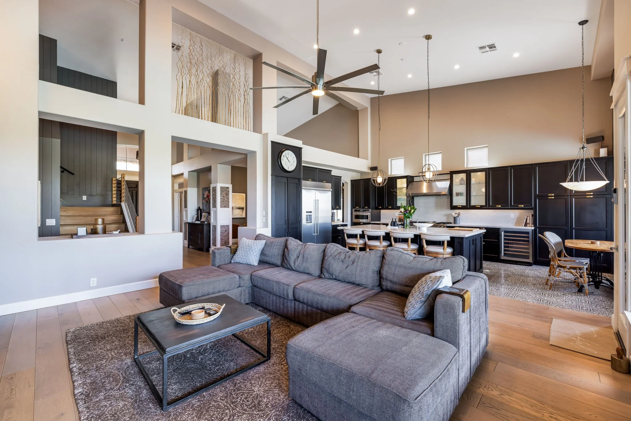 Open-concept living room and kitchen with high ceilings, large gray sectional sofa, black cabinetry, island with bar stools, and modern lighting fixtures.