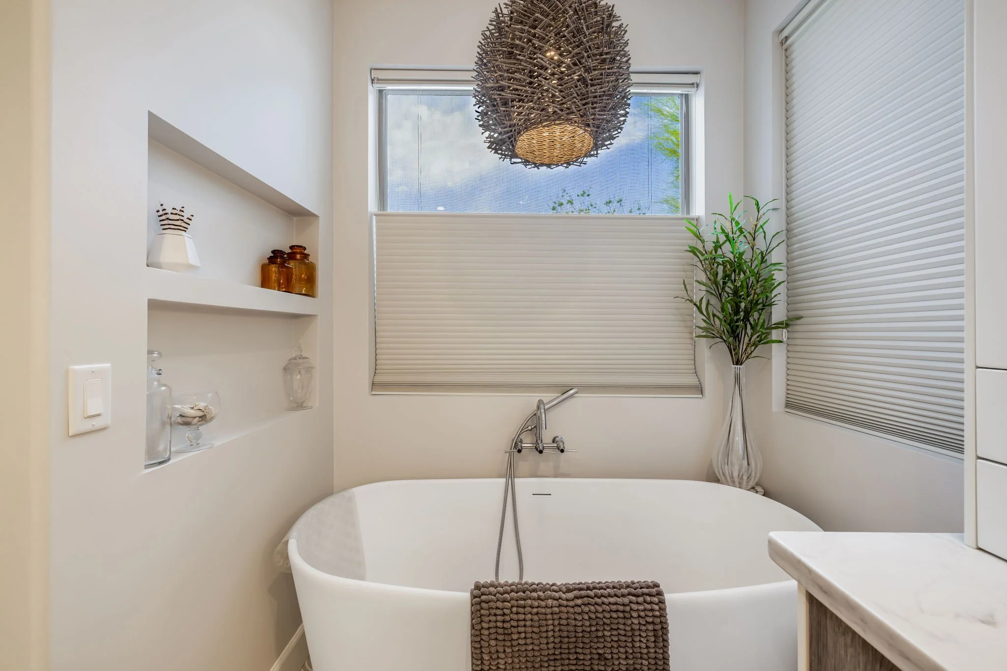Modern bathroom with a white bathtub, a vases with a tall green plant, and built-in shelving with amber and glass bottles, lit by a large woven pendant light.