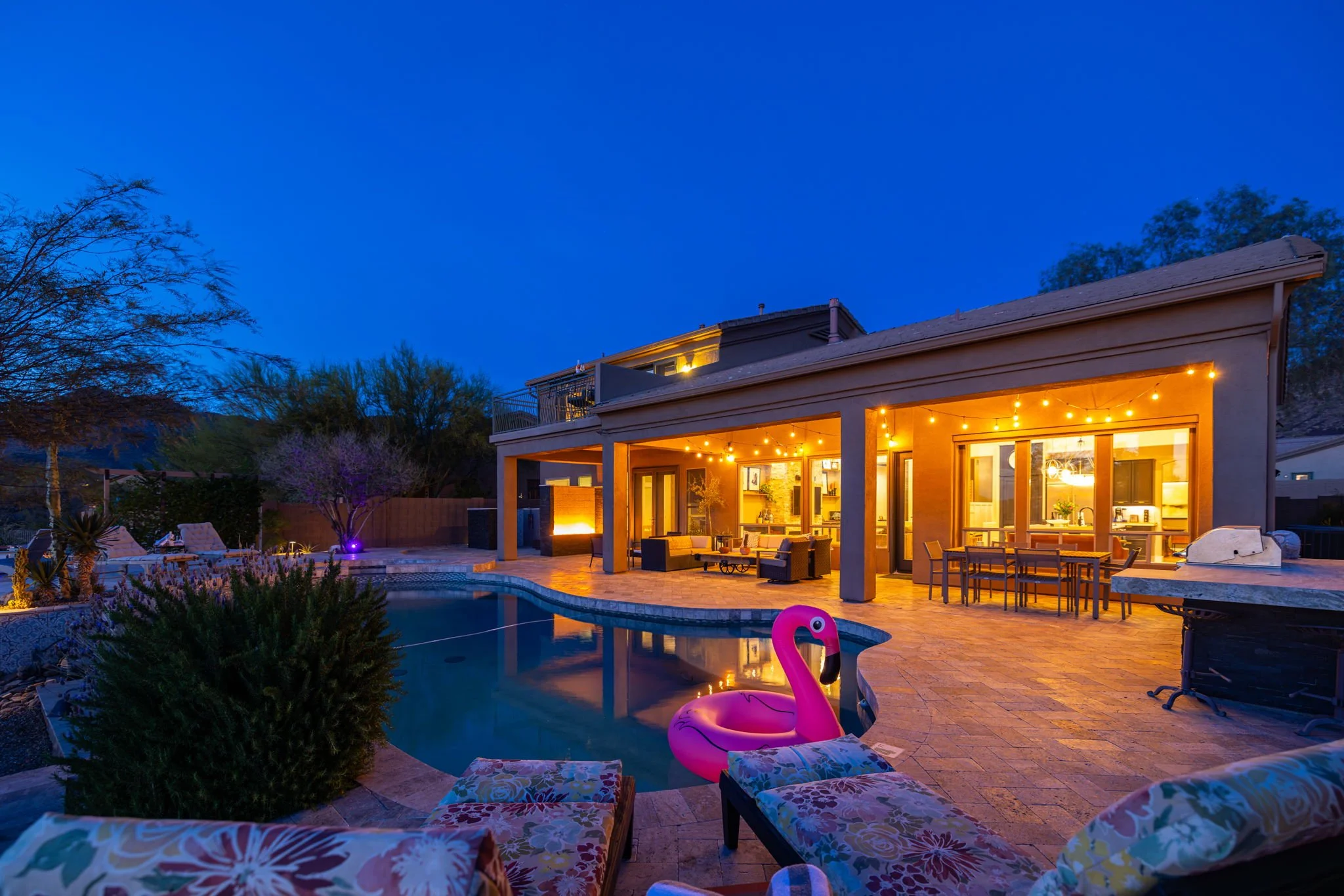 Backyard view of a house at dusk with a lit outdoor patio, swimming pool with an inflatable pink flamingo, lounge chairs, outdoor dining table, and string lights