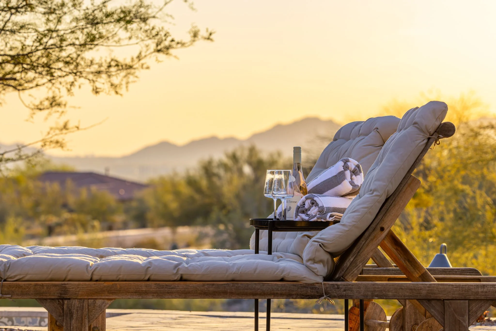 Outdoor lounge chair with a cushion, topped with a tray holding wine, glasses, a bottle, and rolled towels, set in a sunny outdoor area with trees and mountains in the background at sunset.