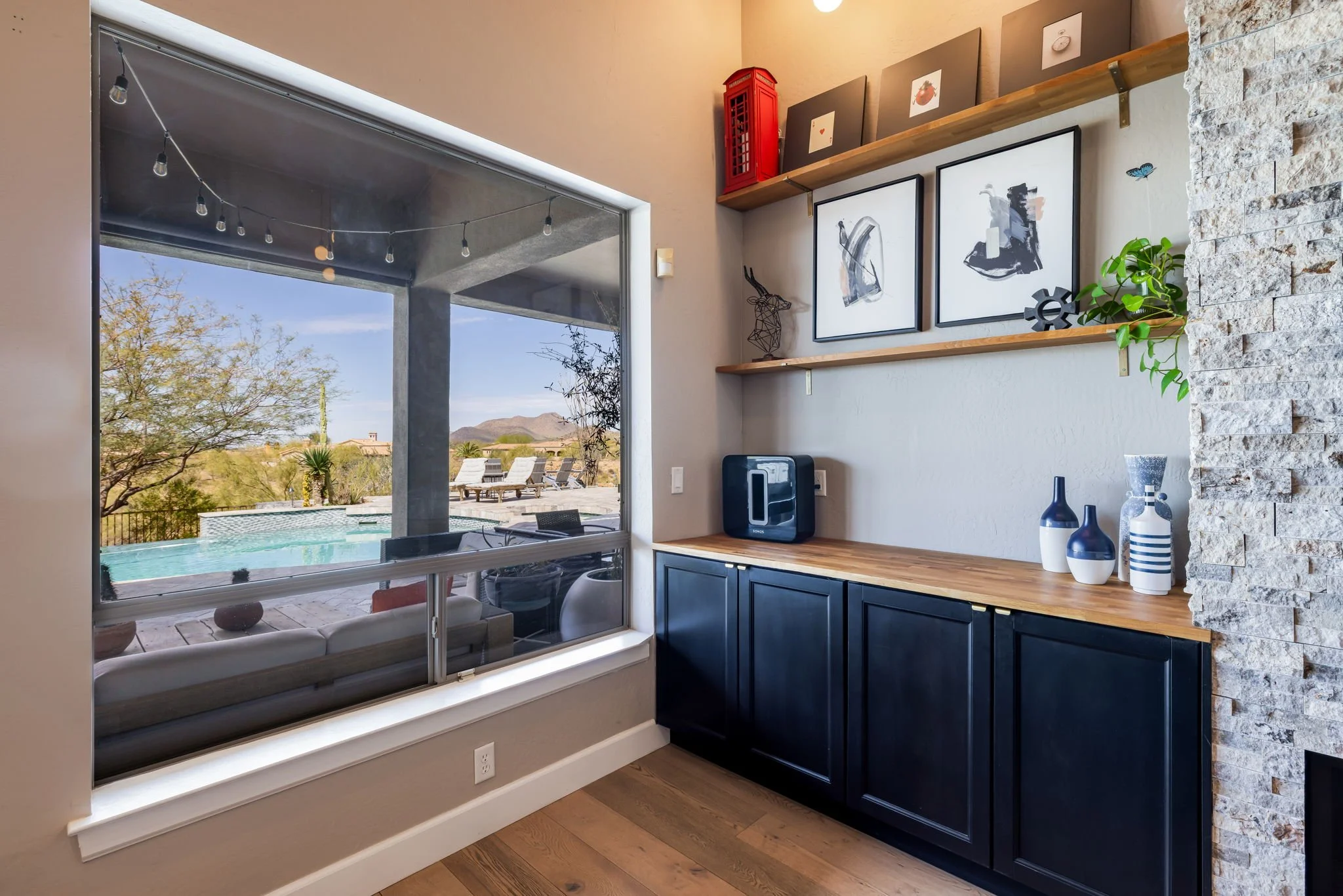 Interior view of a room with a large window revealing a backyard with a swimming pool, outdoor chairs, and desert landscape with mountains in the background. Inside, there are black cabinets with a wooden countertop, framed pictures hanging on the wa