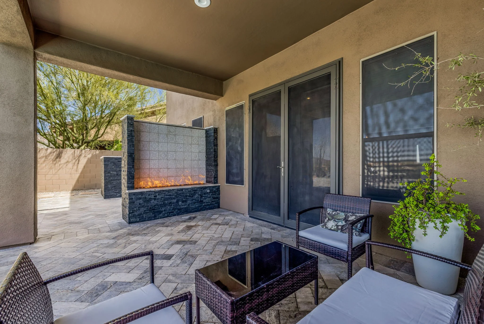Patio area with outdoor furniture, a large potted plant, a wall fountain with orange flames, and sliding glass doors.