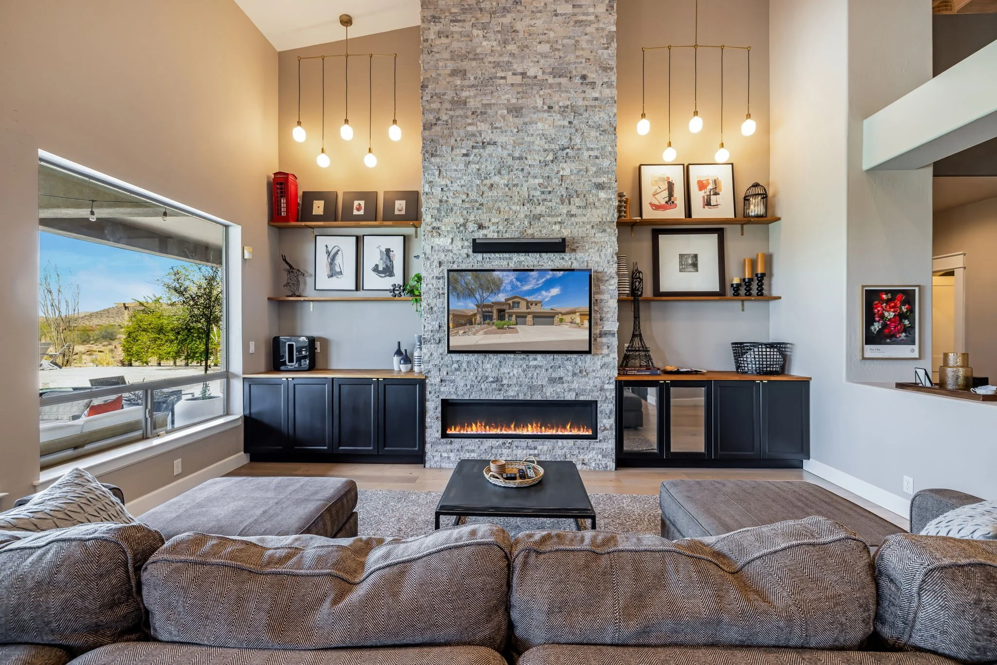 Living room with large window, modern fireplace, and built-in shelves with wall art and decor, overlooking a desert landscape.