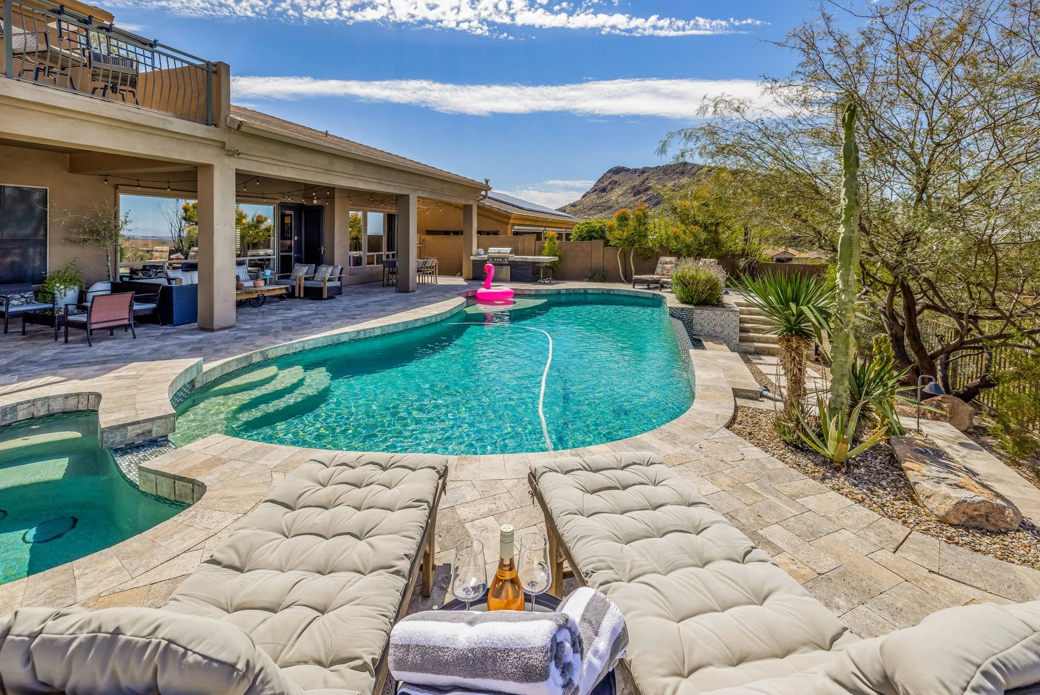 Backyard with a kidney-shaped swimming pool, beige lounge chairs, a pool toy swan, a bottle of rosé, glasses, and towel, surrounded by desert plants and a house with a covered patio, with a mountain in the background.