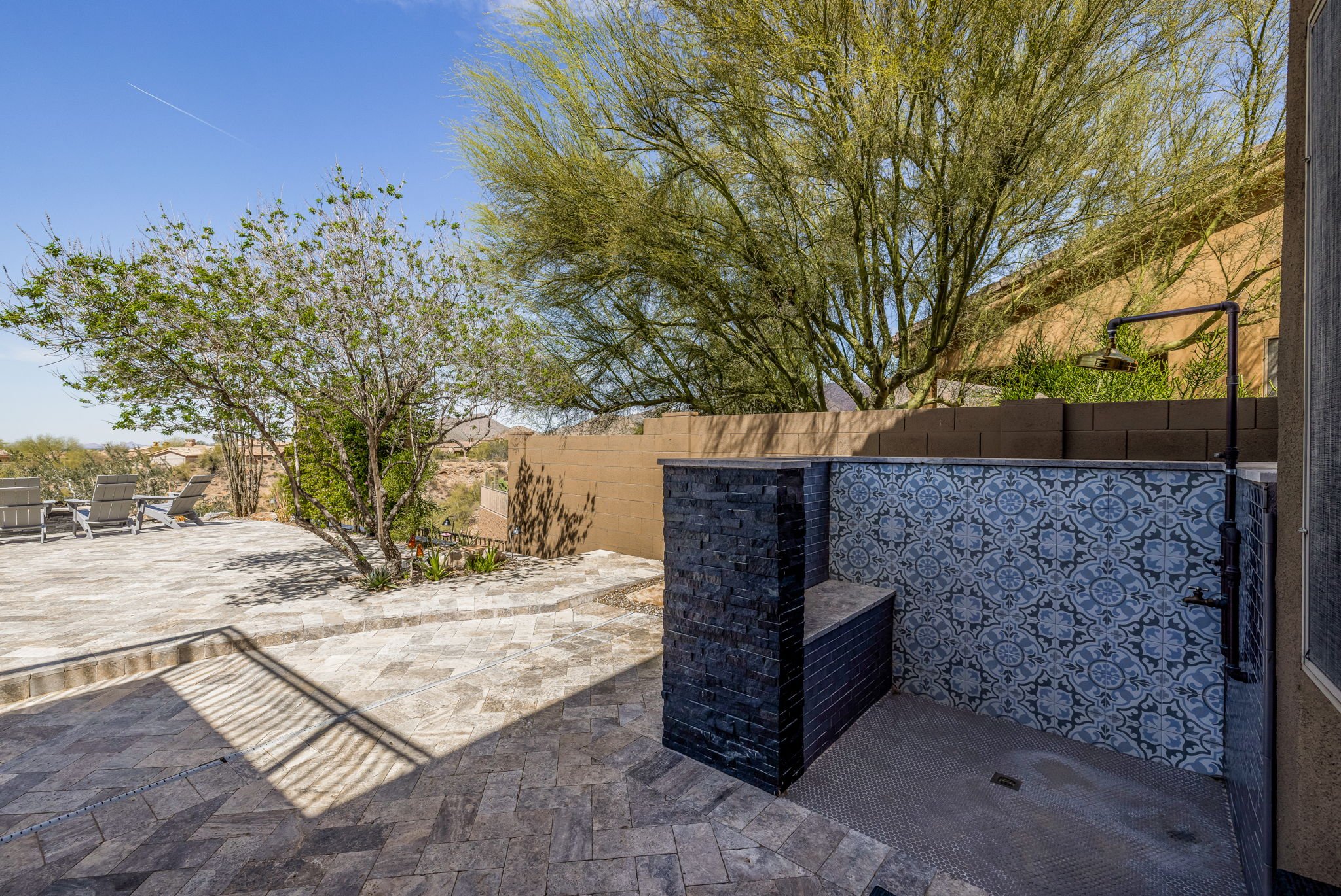 Outdoor patio area with stone flooring, trees, and lounge chairs, featuring an outdoor shower with blue patterned tiles.