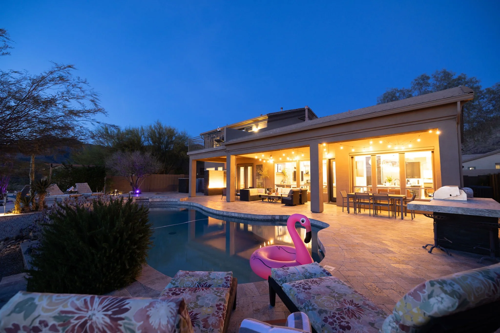 A backyard pool area at dusk with outdoor furniture, string lights, a pink flamingo pool float, and a view of the house with illuminated interior.