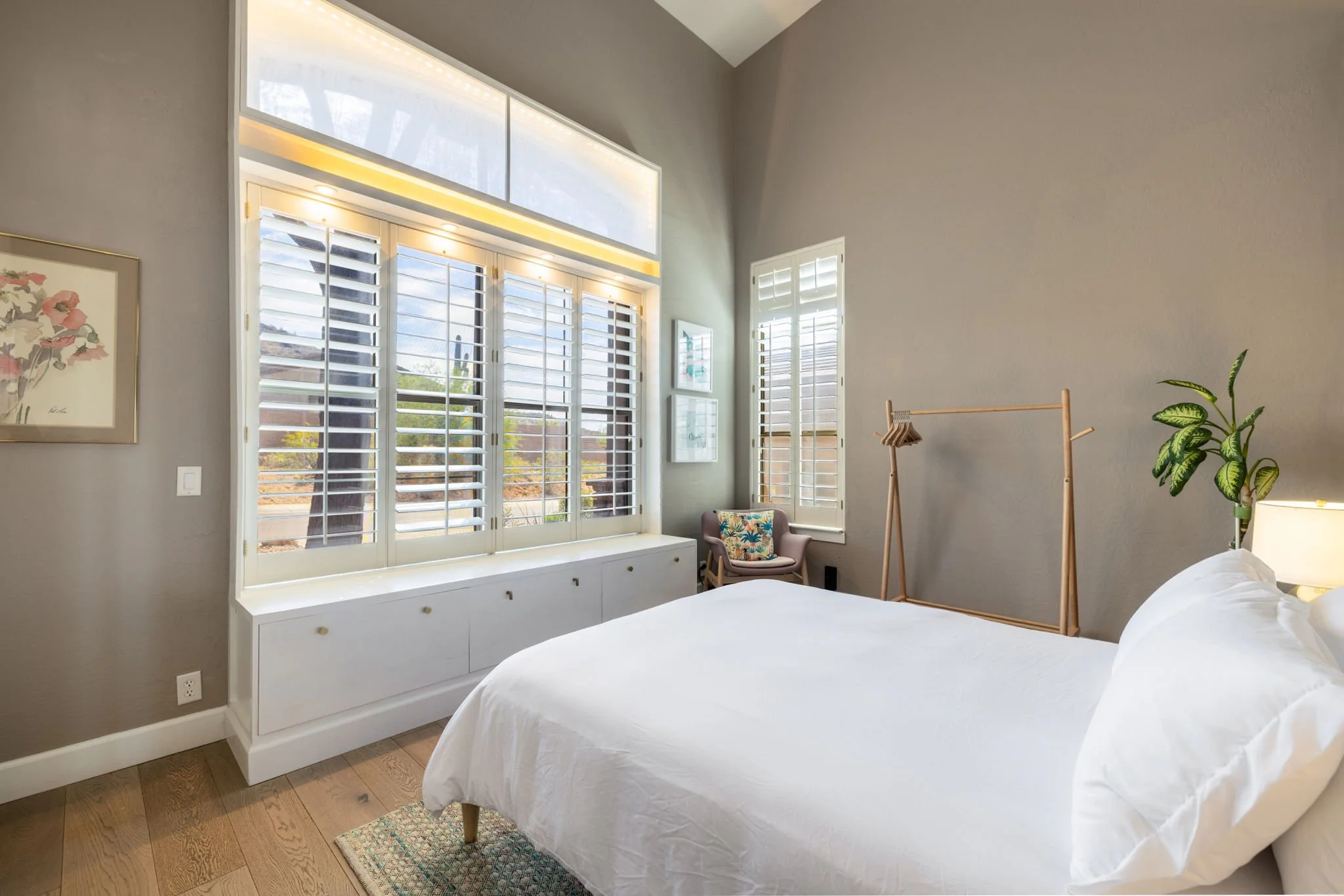 A bedroom with a large window with white shutters, a white bed, a small armchair with a colorful pillow, a wooden clothing rack, a potted plant, and framed artwork on the wall.