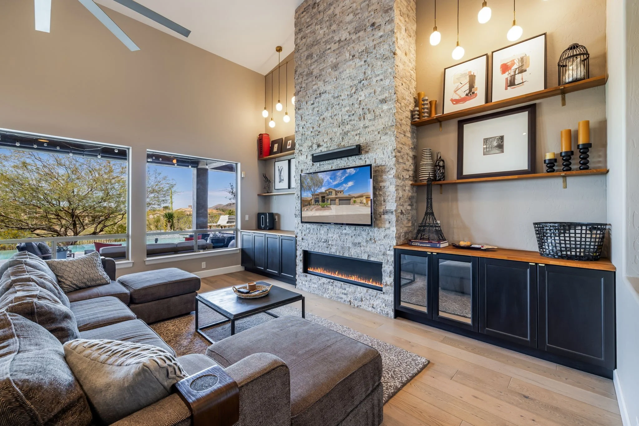 Living room with a sectional sofa, a wall-mounted TV above a fireplace, large windows with a view of trees outside, and shelves with decor and artwork.