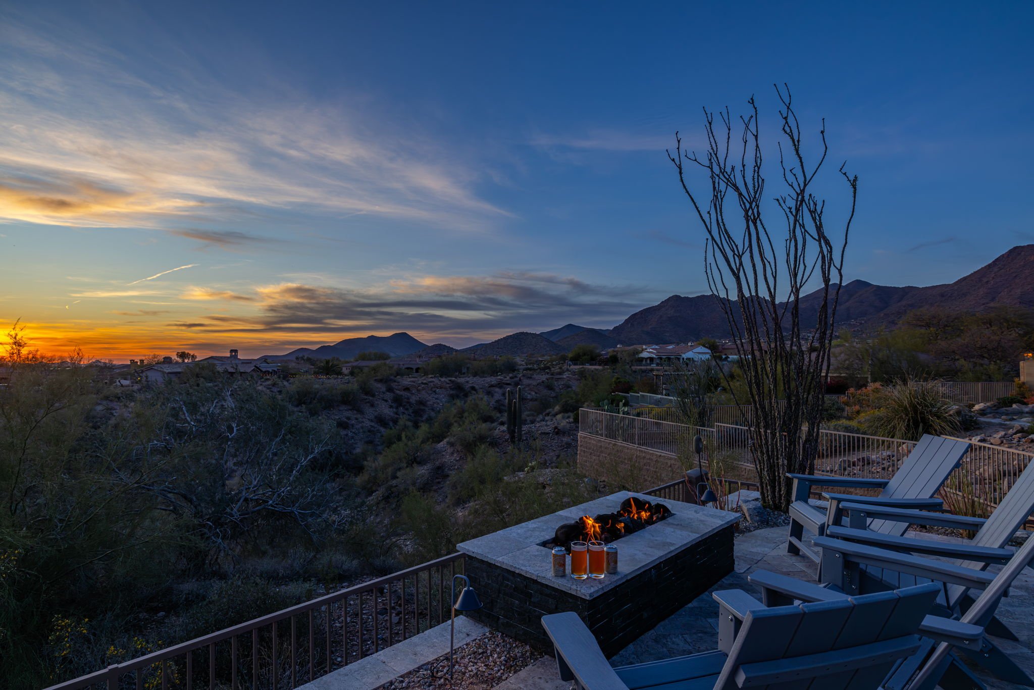 Outdoor patio with chairs and a fire pit, overlooking a desert landscape and mountains at sunset.
