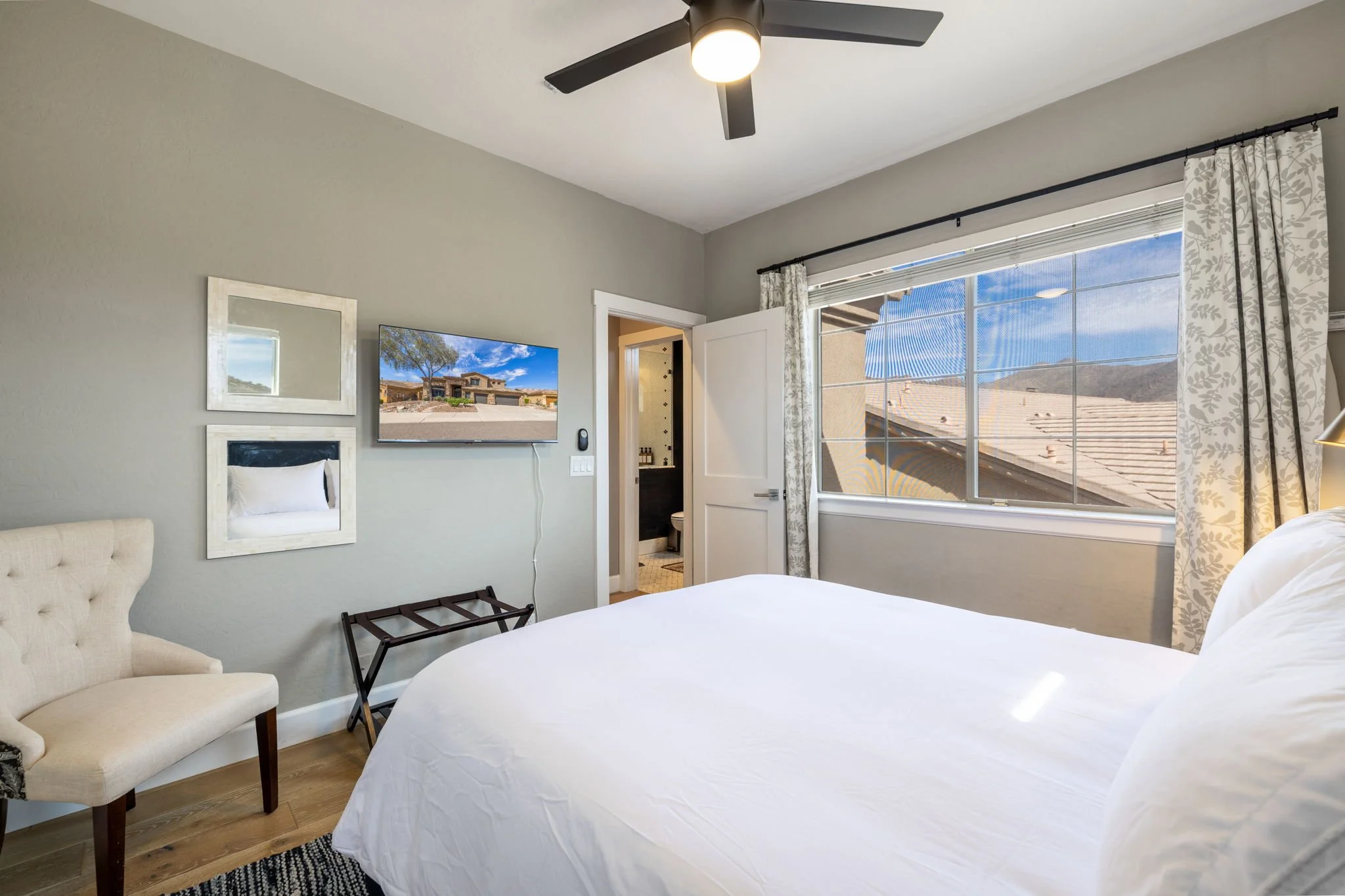 Bright hotel bedroom with large window, white bed, beige armchair, wall-mounted TV, and an adjoining bathroom.