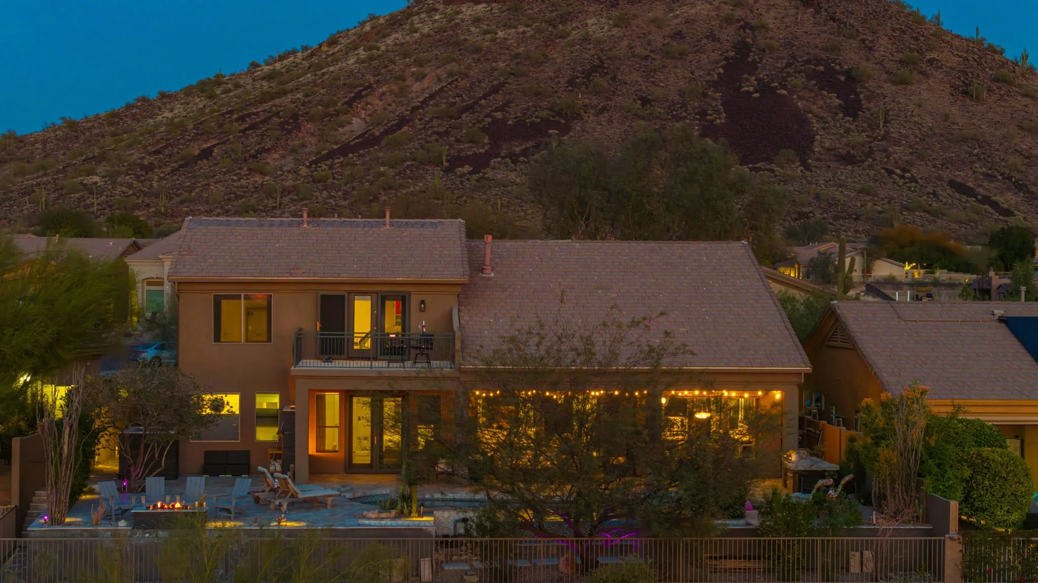 Nighttime view of a two-story house with warm lighting, a backyard patio with lounge chairs and a fire pit, surrounded by desert plants and trees, with a mountain in the background.