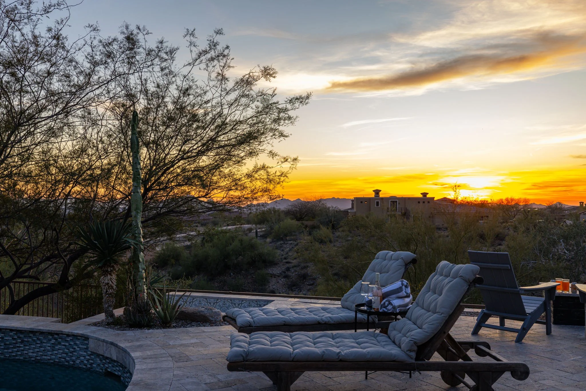Outdoor patio area at sunset with lounge chairs, a small table with bottles and glasses, a tree, desert bushes, and a distant house against silhouettes of mountains.