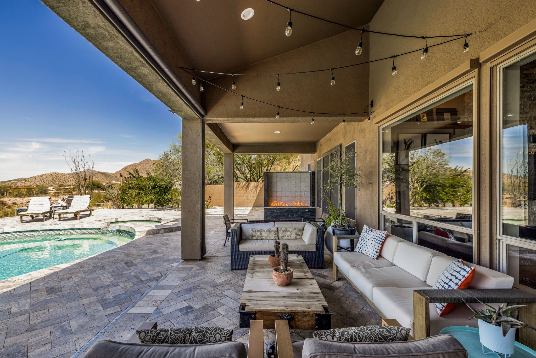 Outdoor patio with seating area, sofas with pillows, and a fire feature, overlooking a backyard with a pool and lounge chairs, in a desert landscape with mountains in the background, under string lights.