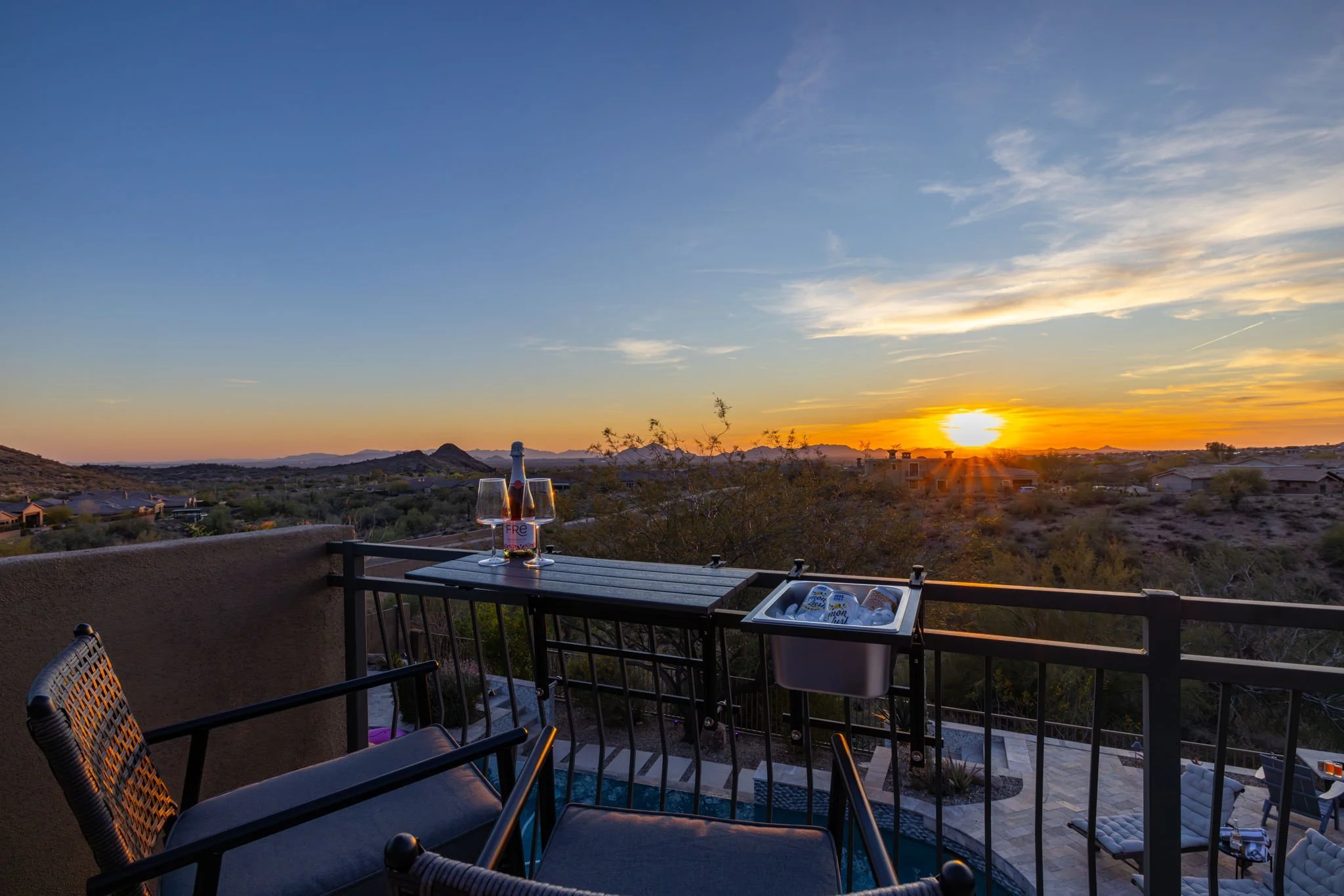 Balcony with a table, chairs, a bottle of sparkling wine and glasses, overlooking a desert landscape at sunset with houses and mountains in the distance.