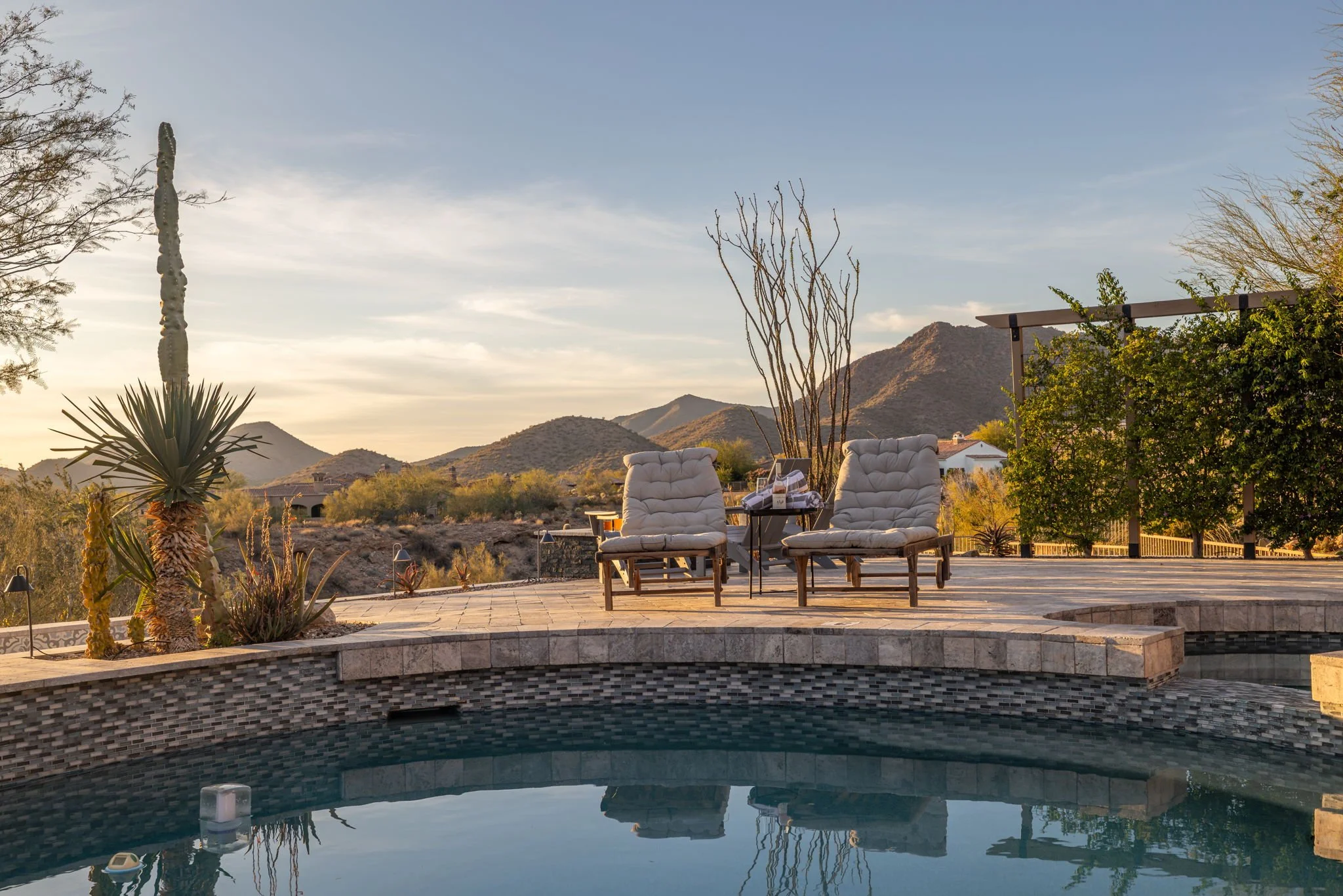 Arizona desert landscape with mountains, a swimming pool, two cushioned outdoor chairs, and plants including cactus and trees, during sunset.