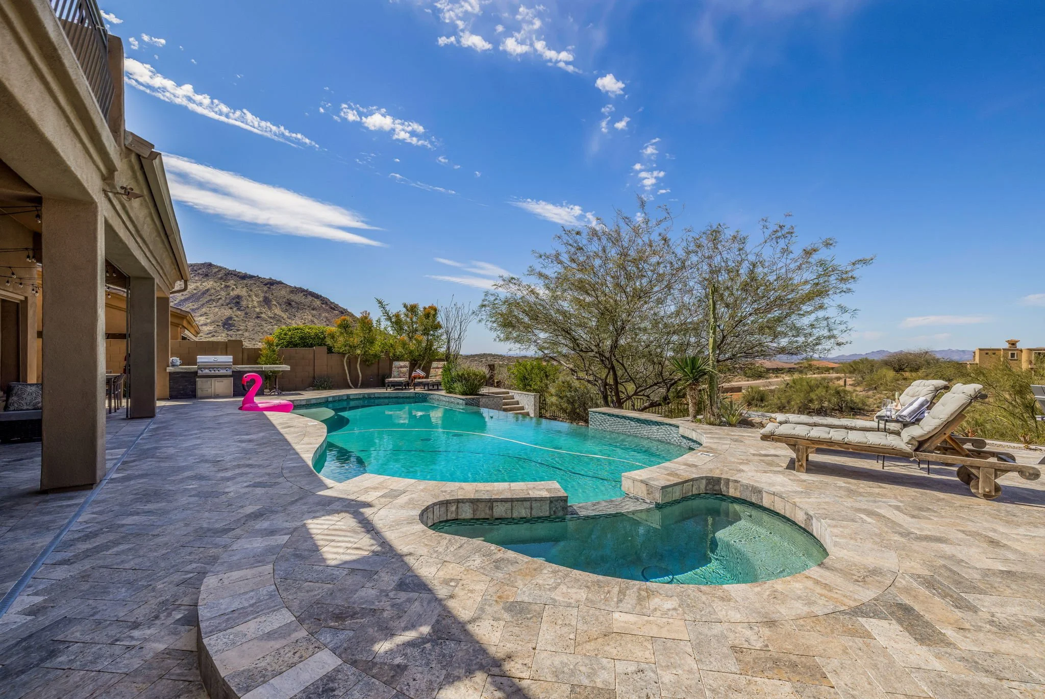 Backyard with a swimming pool, hot tub, lounge chairs, and a desert landscape with mountains in the background under a partly cloudy sky.