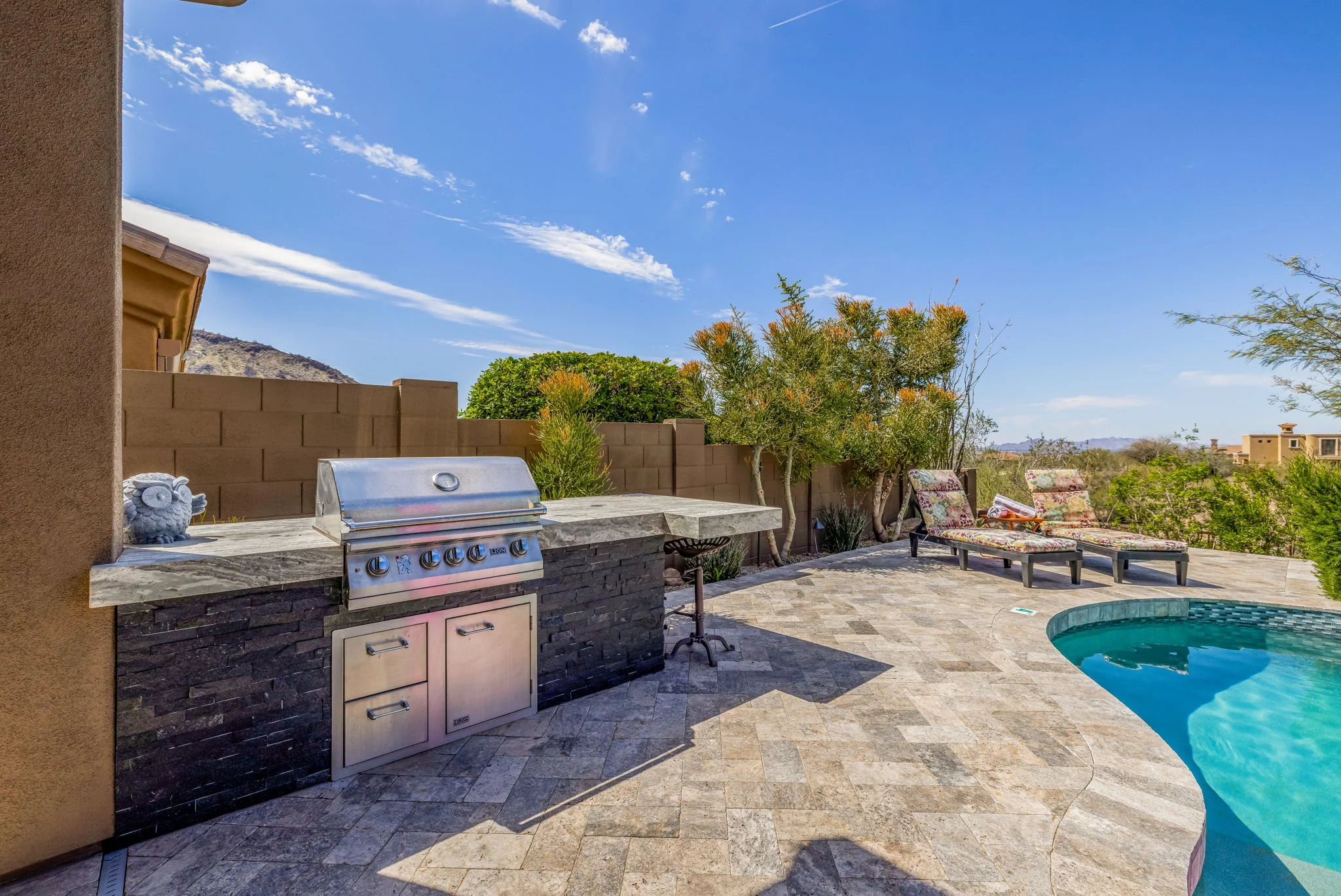 A backyard with a built-in barbecue grill, two lounge chairs, and a pool, with desert landscaping and mountain views under a blue sky.