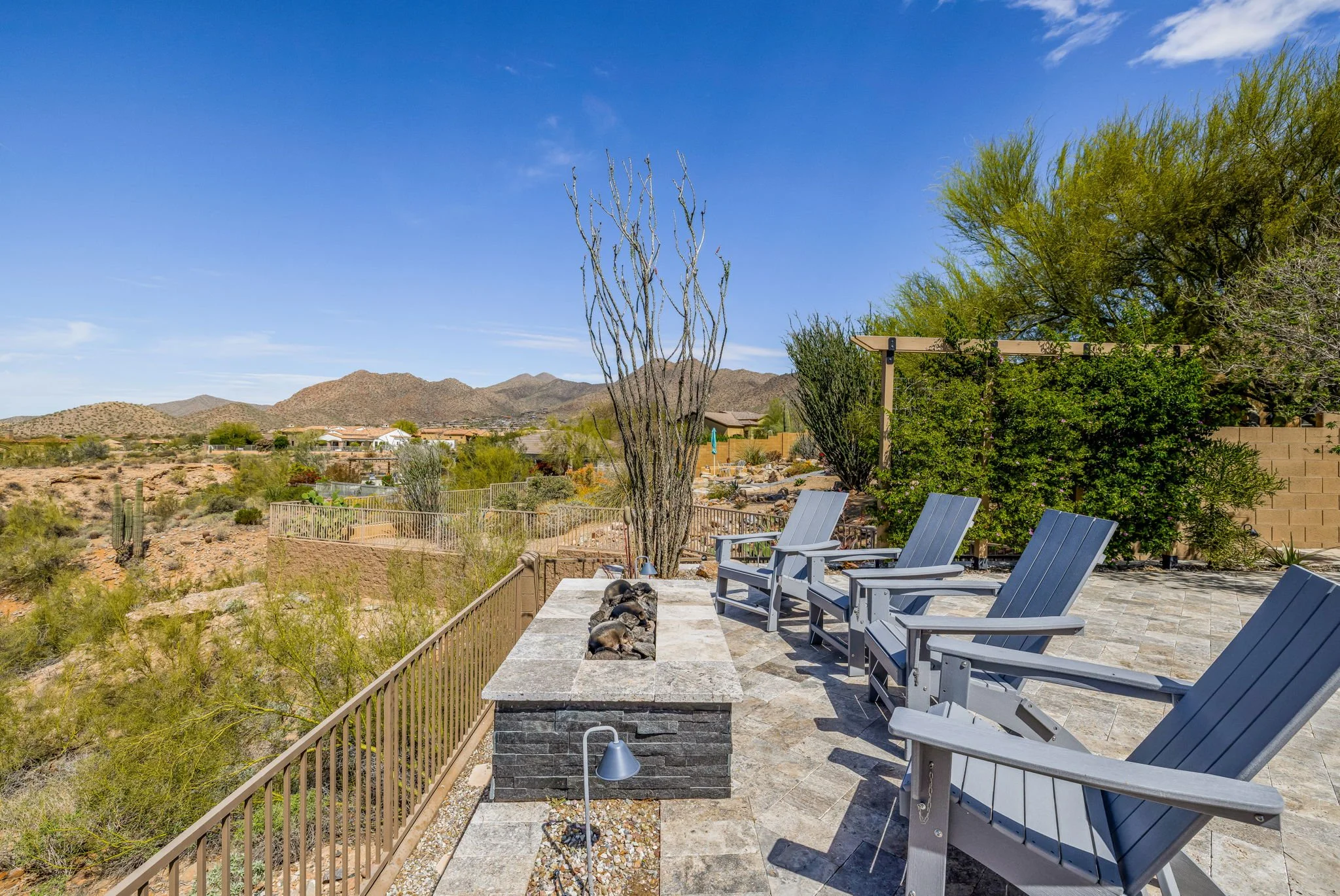 Desert backyard patio with four blue Adirondack chairs, a built-in fire pit with a dog sleeping on it, desert plants, cacti, and mountain landscape in the background under a blue sky.