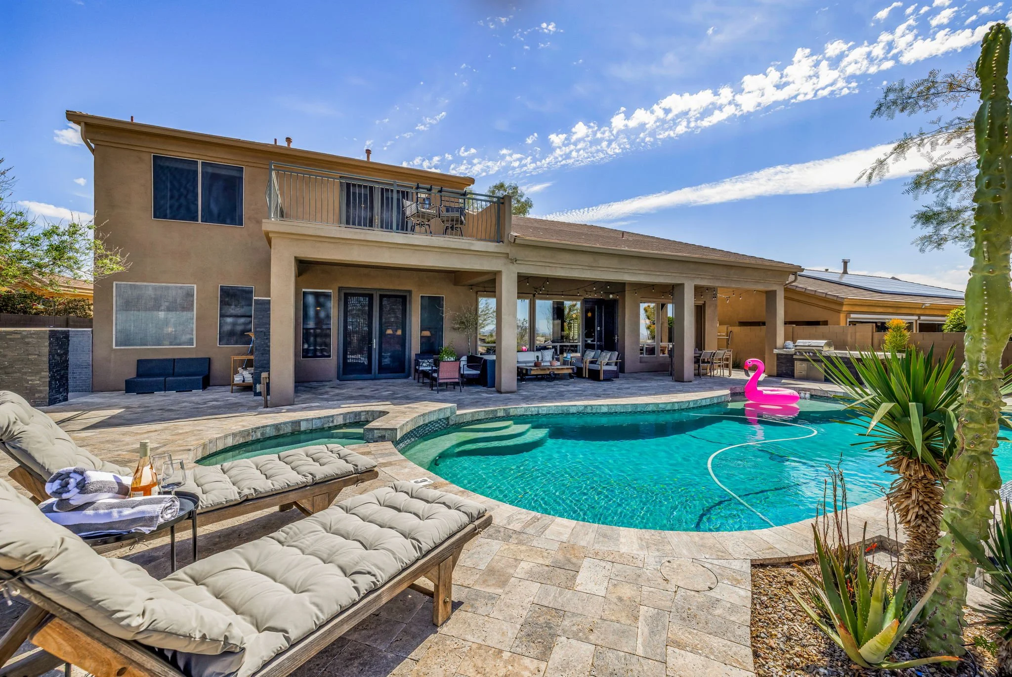 Backyard with a swimming pool, lounge chairs, patio furniture, a pink flamingo float, a barbecue grill, and a two-story house with a balcony and sliding glass doors, under a blue sky with clouds.