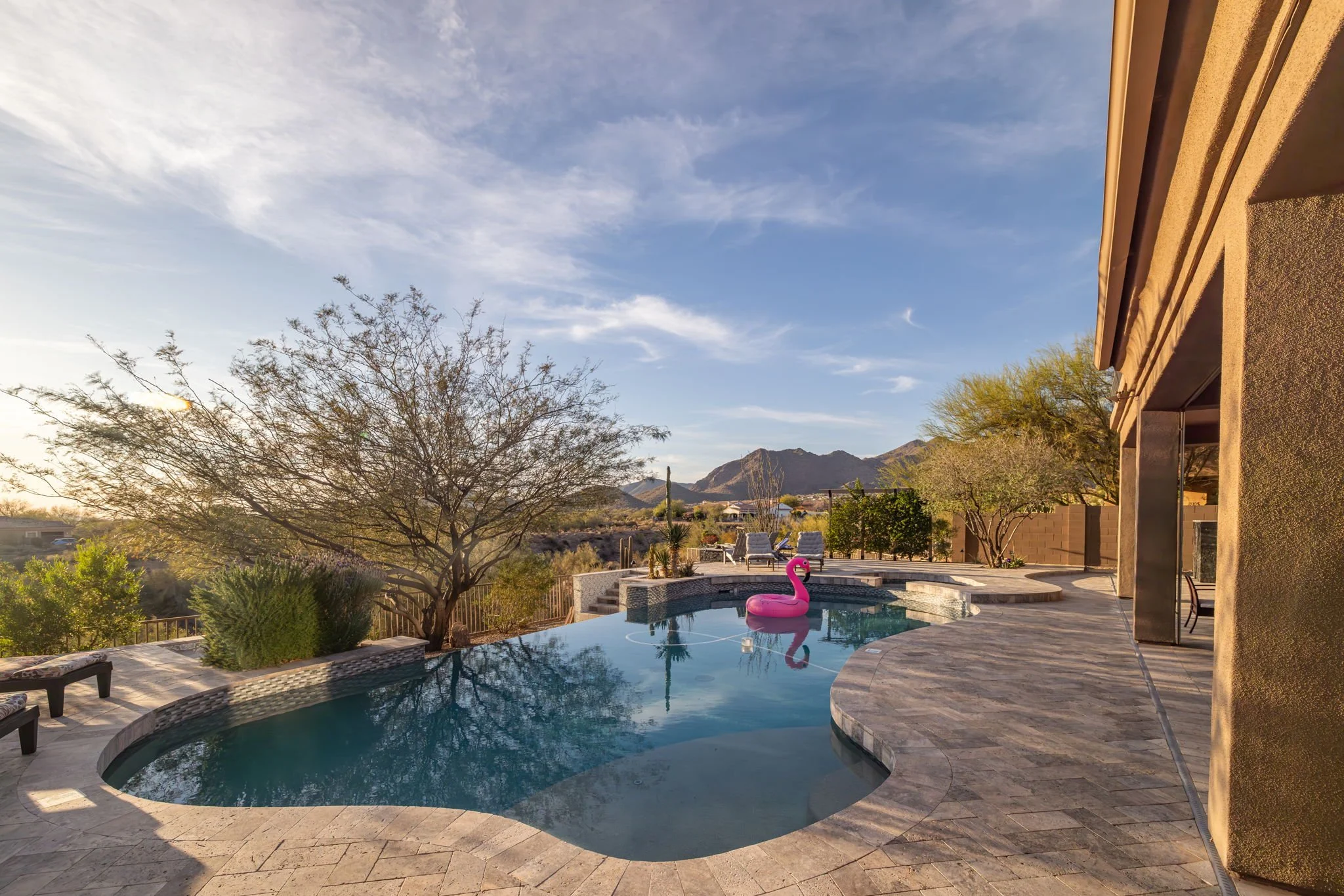 A backyard swimming pool with a pink flamingo float, surrounded by desert landscape and mountains in the background, under a partly cloudy sky.
