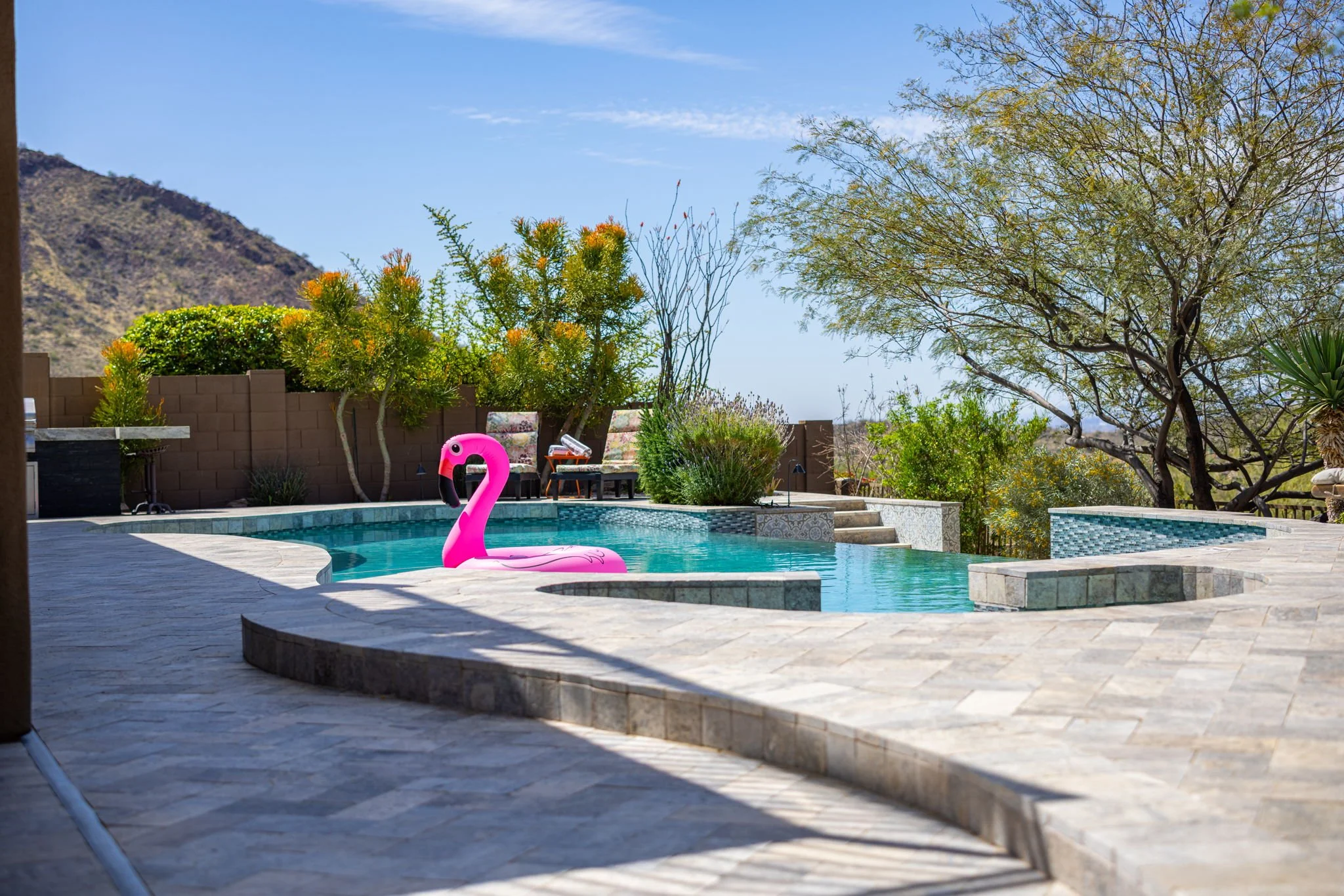 Backyard swimming pool with a pink flamingo float, surrounded by trees and patio furniture under a clear blue sky.