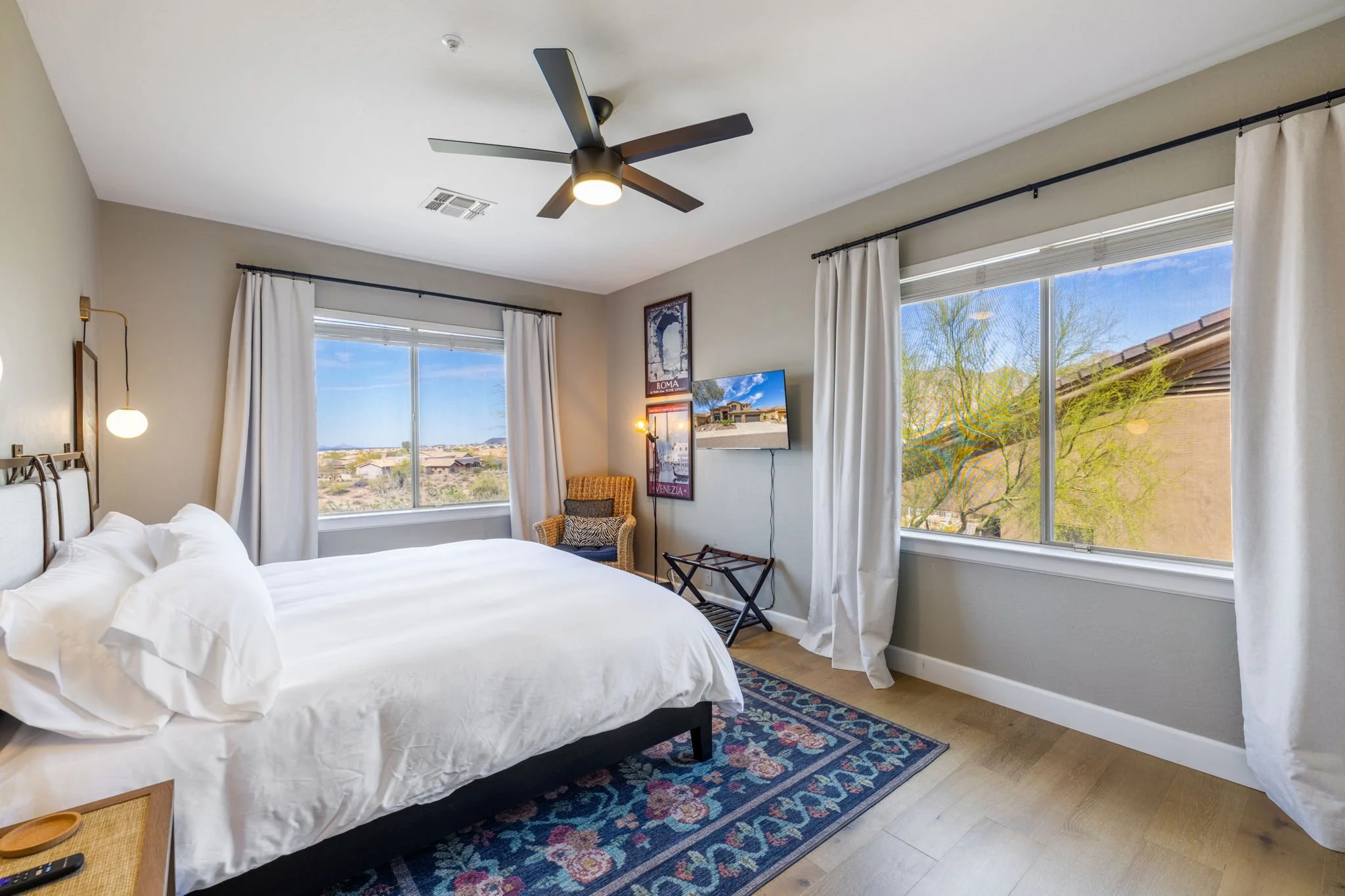 Hotel room with a bed, windows with white curtains showing a view of a desert landscape, a rug, a wall-mounted TV, a chair, and modern lighting.