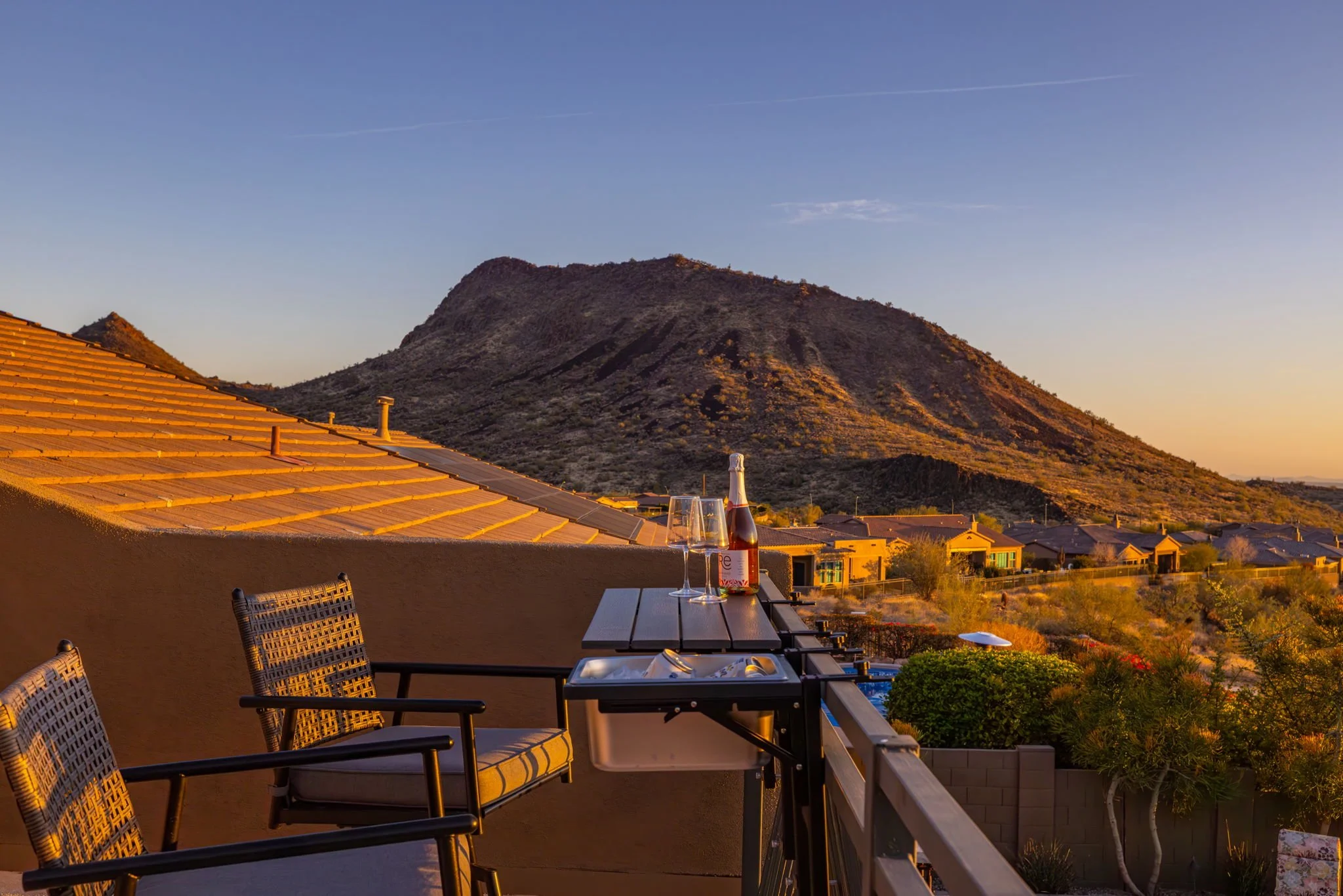 Sunset view from a balcony with two chairs, a small table with a bottle of wine and two wine glasses, overlooking a neighborhood with mountains in the background.